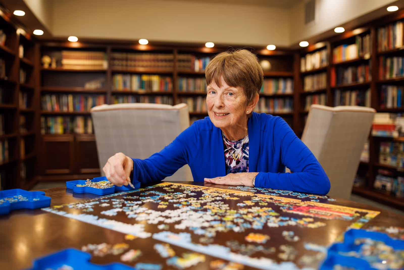 An elderly woman wearing a blue cardigan is sitting at a table working on a jigsaw puzzle in a room with bookshelves filled with books in the background.