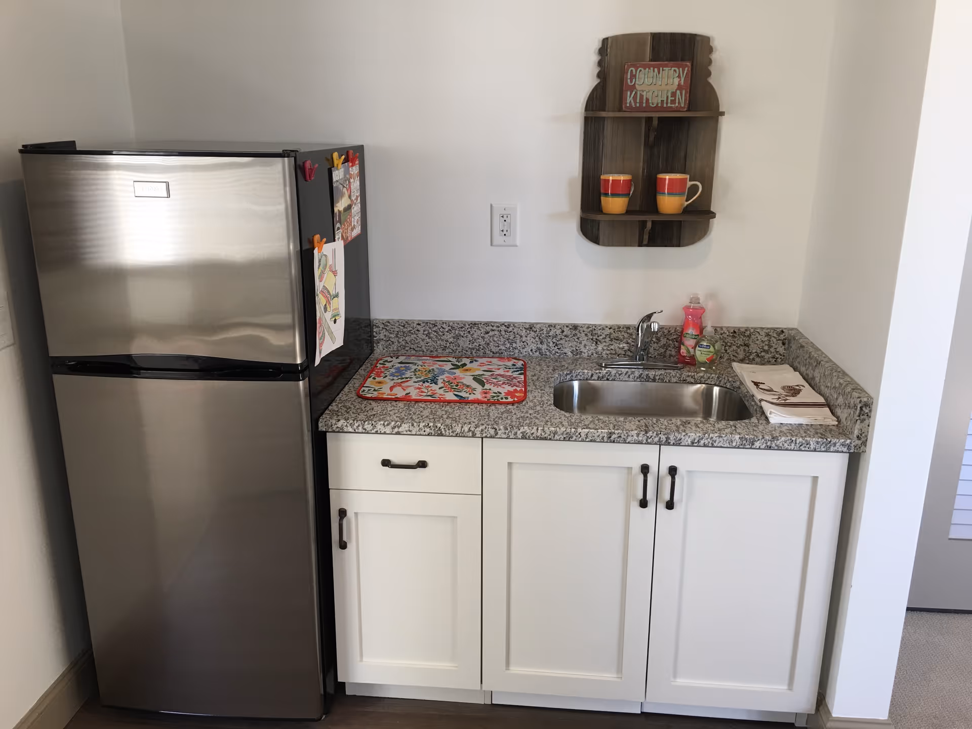 Small kitchenette with a stainless fridge, granite counter, sink, white cabinets, and a decorative shelf.