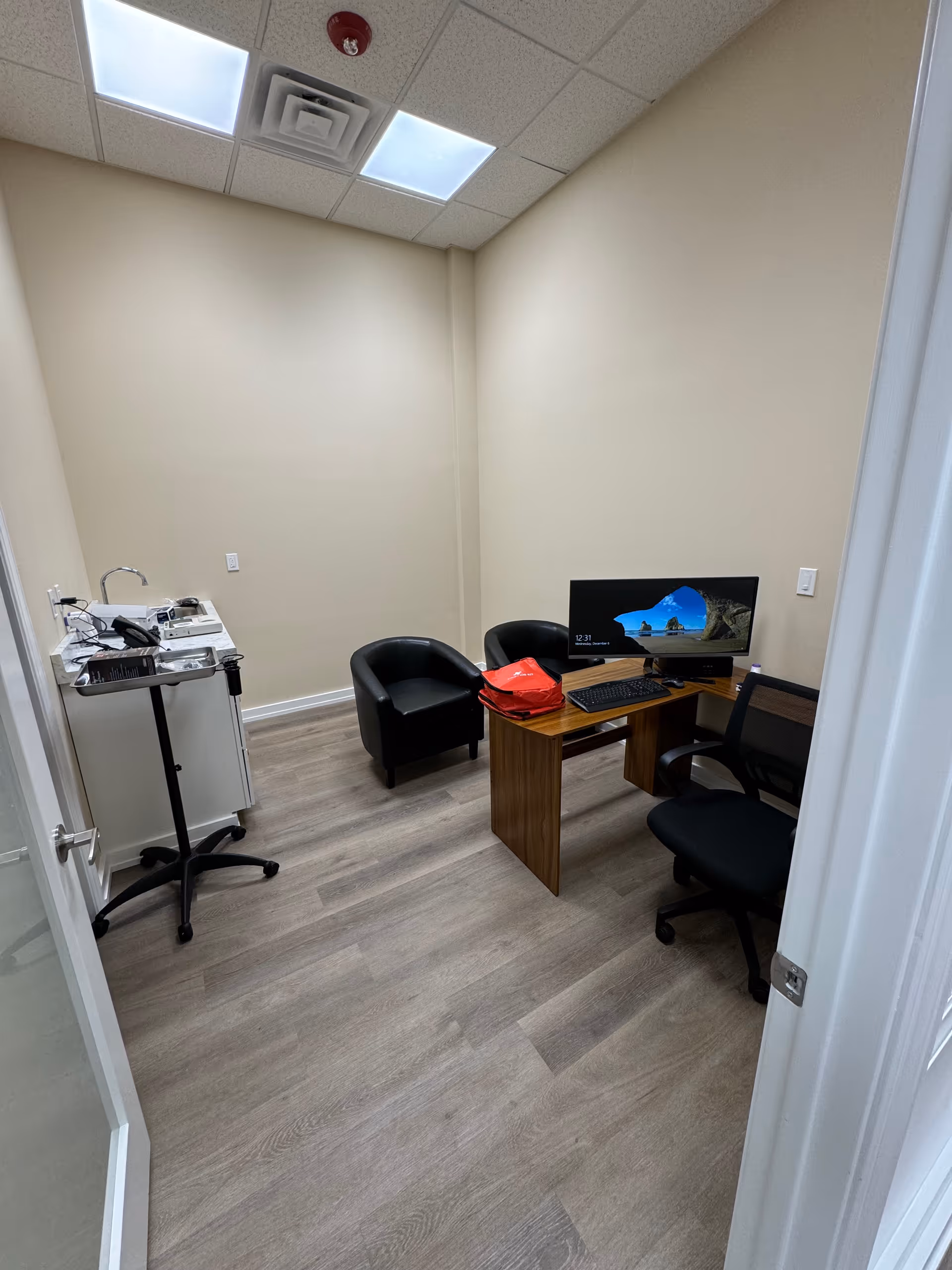 Small clinic-like office with desk, computer, chair, and a black armchair.