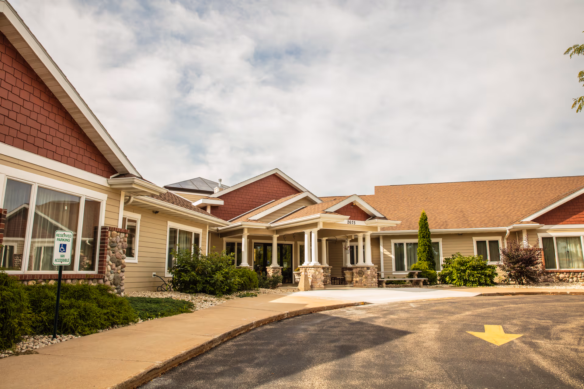Exterior view of Oakwood Manor senior living facility showing a single-story building with beige siding, red shingle accents, stone pillars, and a covered entrance. There is a reserved parking sign for handicapped accessibility near the sidewalk and a yellow directional arrow painted on the driveway.