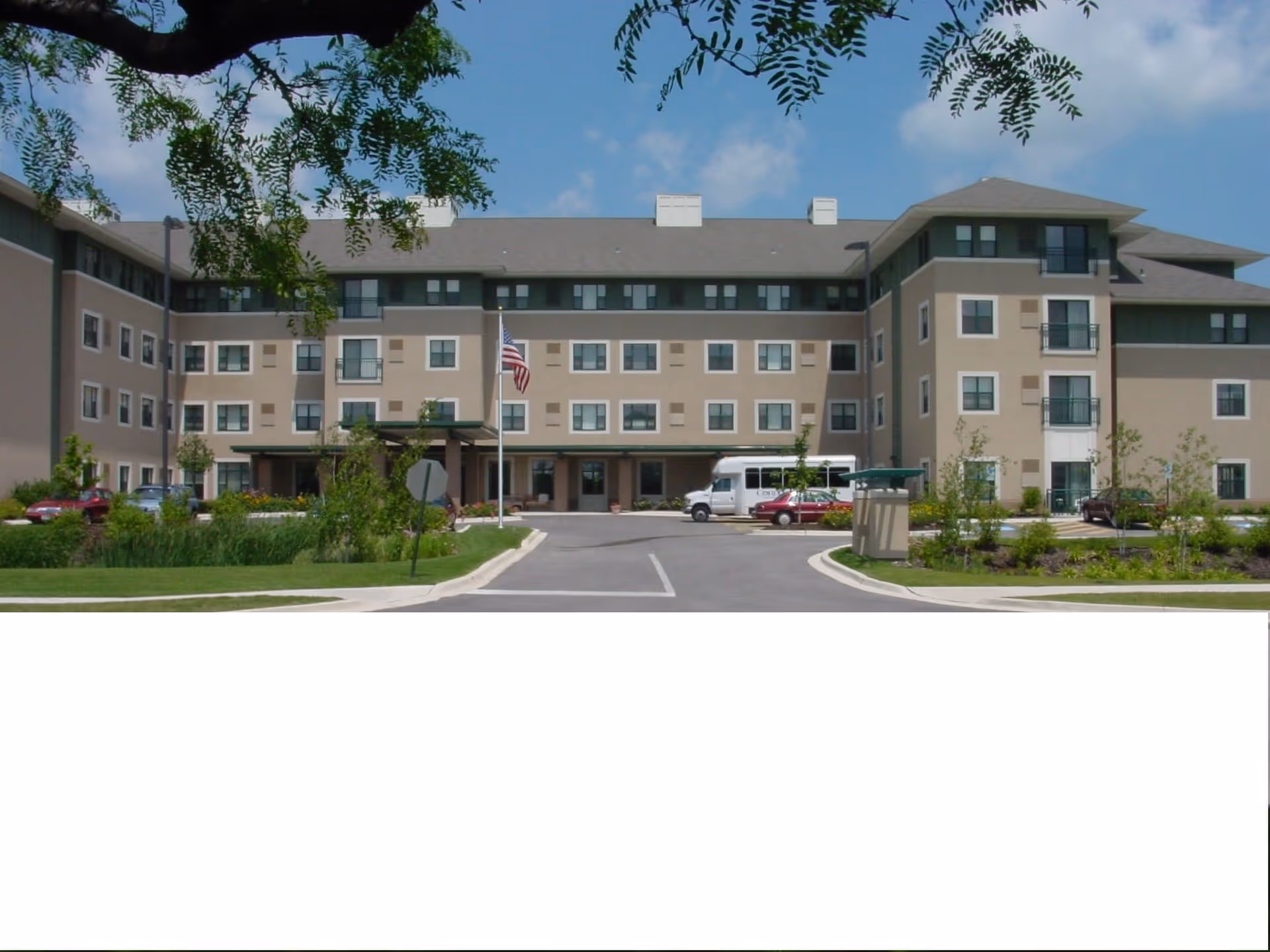 Front exterior view of a multi-story senior living facility building with beige and green walls, multiple windows, an American flag on a flagpole, a driveway with parked cars and a shuttle bus, and landscaped greenery in the foreground under a partly cloudy blue sky.