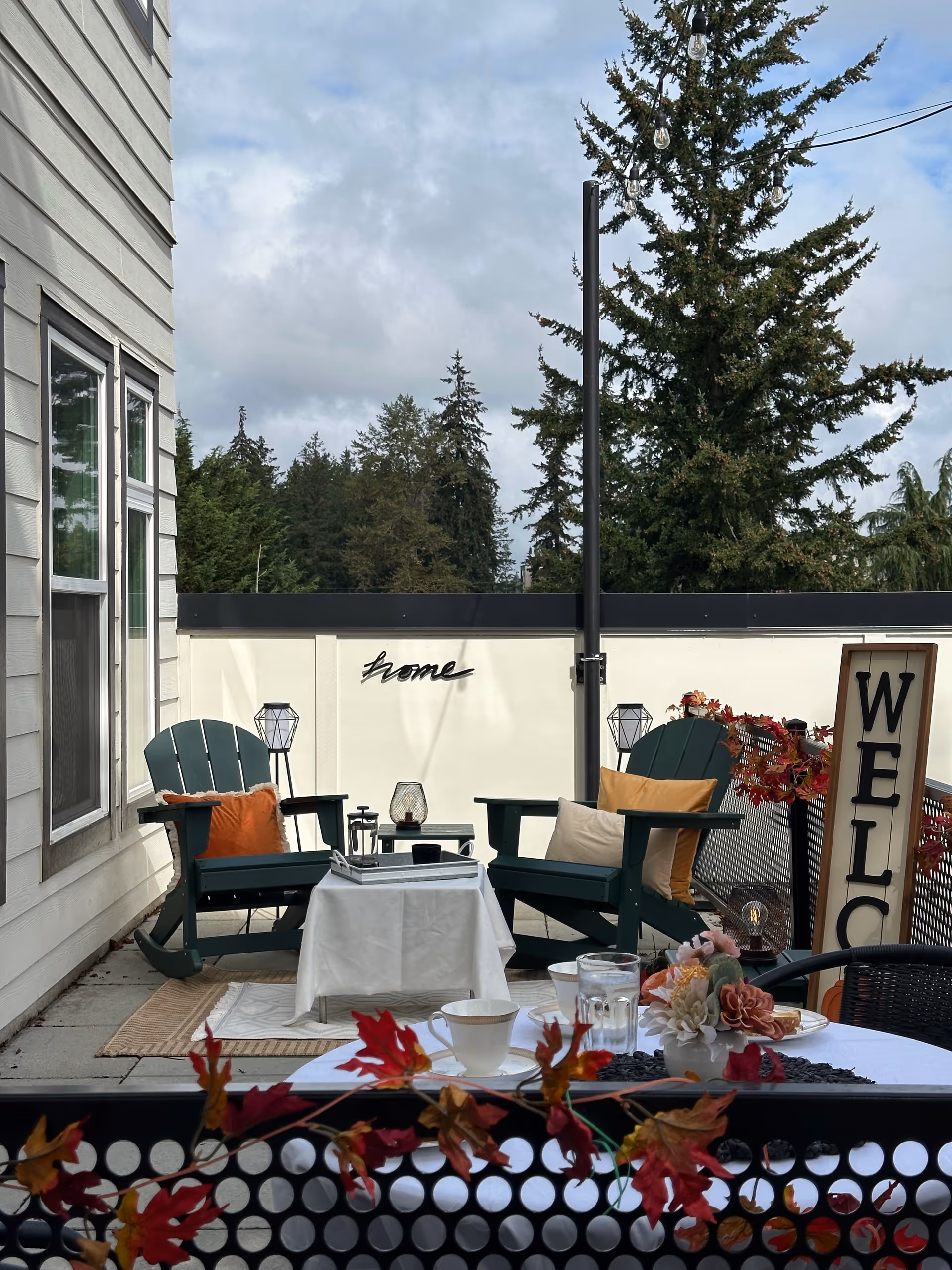 Outdoor patio with two green chairs, table, welcome sign, string lights, and autumn decorations.