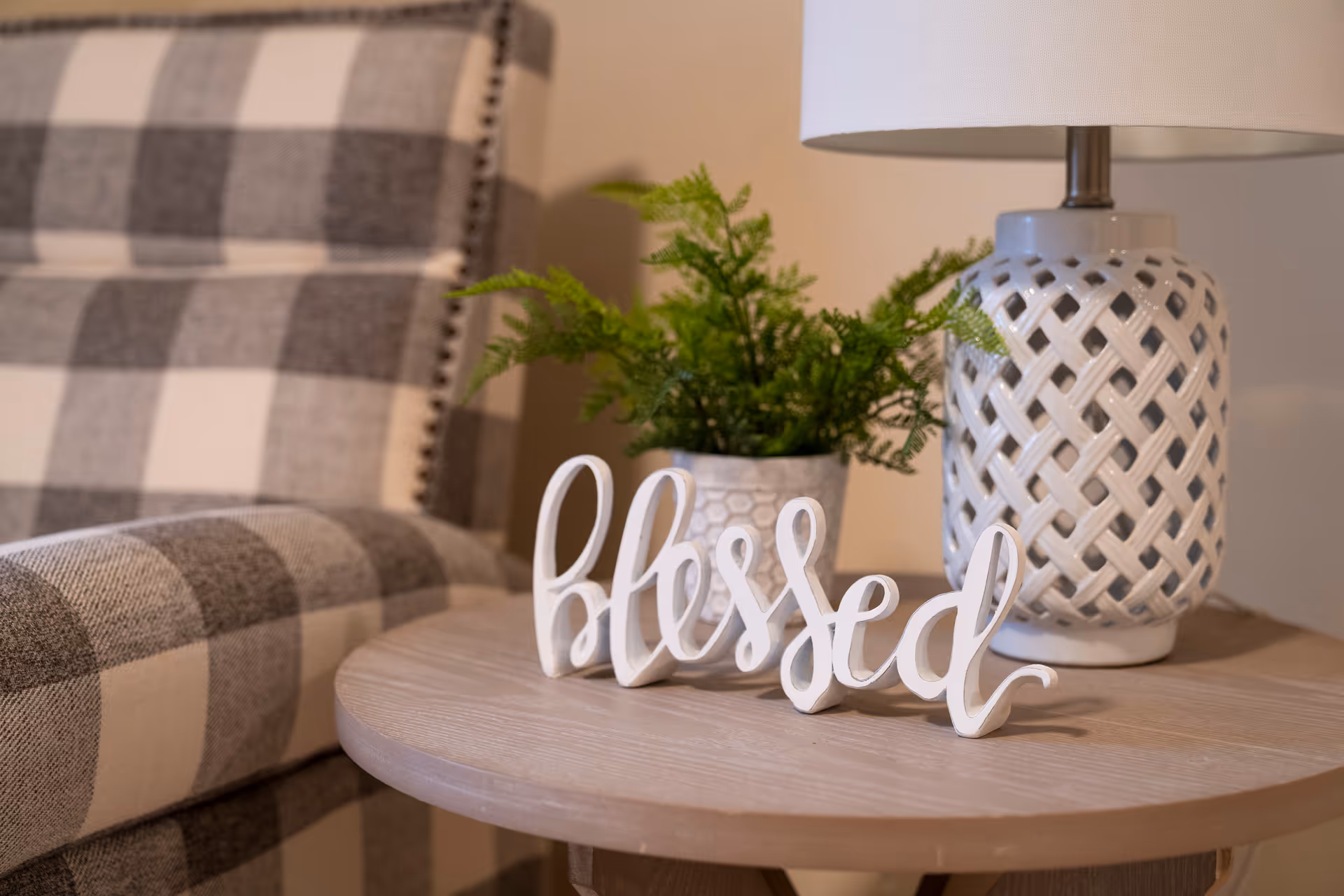 Close-up of a round wooden table with lamp, fern, and 'blessed' decor in a plaid lounge.