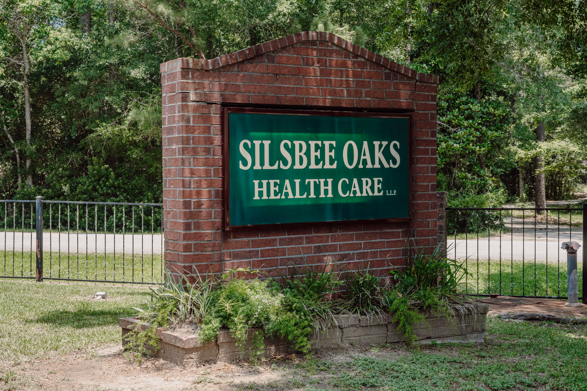 Brick entrance sign that reads 'Silsbee Oaks Health Care' set in a landscaped area with trees and a metal fence.