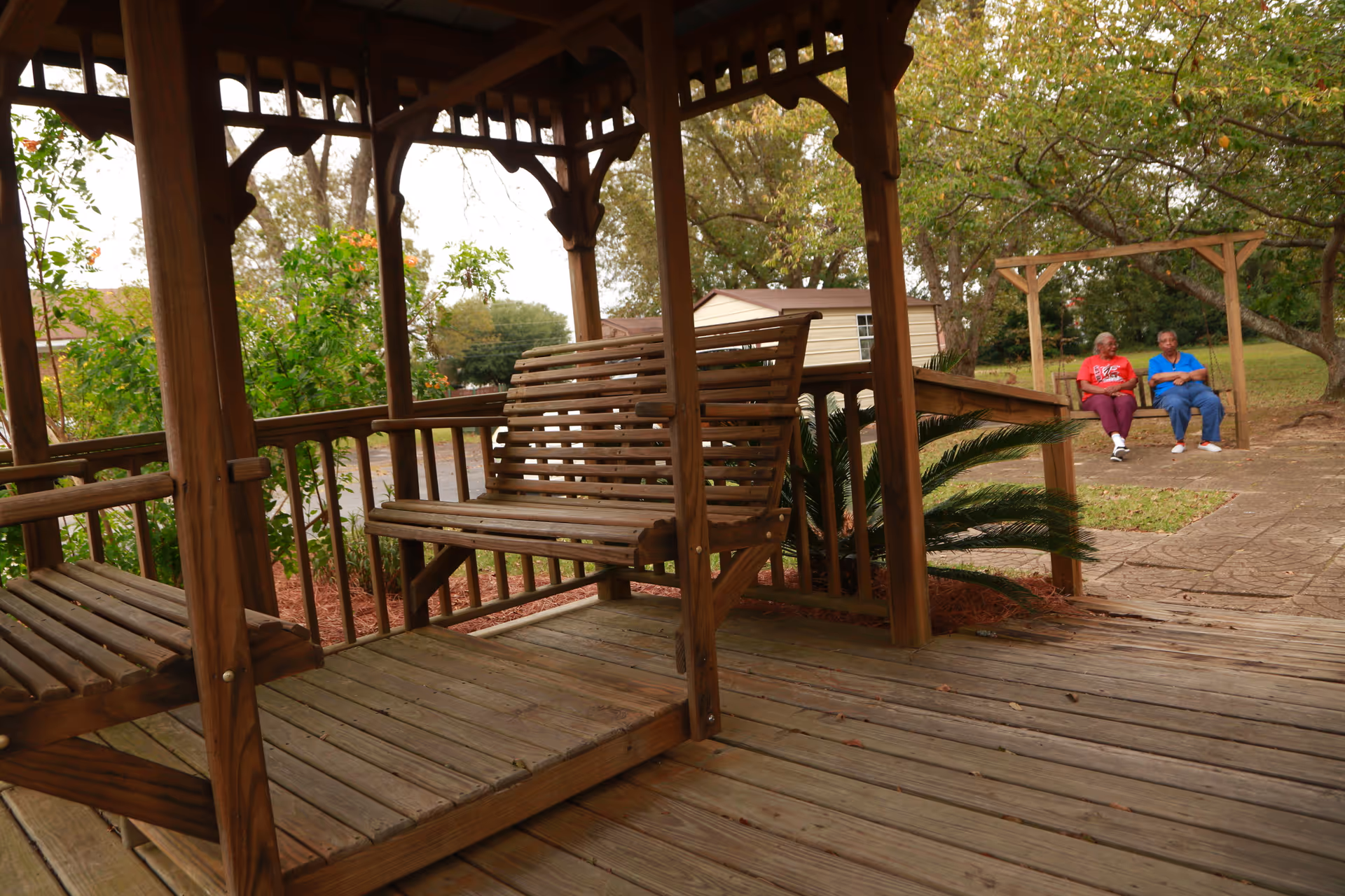 Wooden gazebo with a swing in the foreground and two residents seated outdoors.