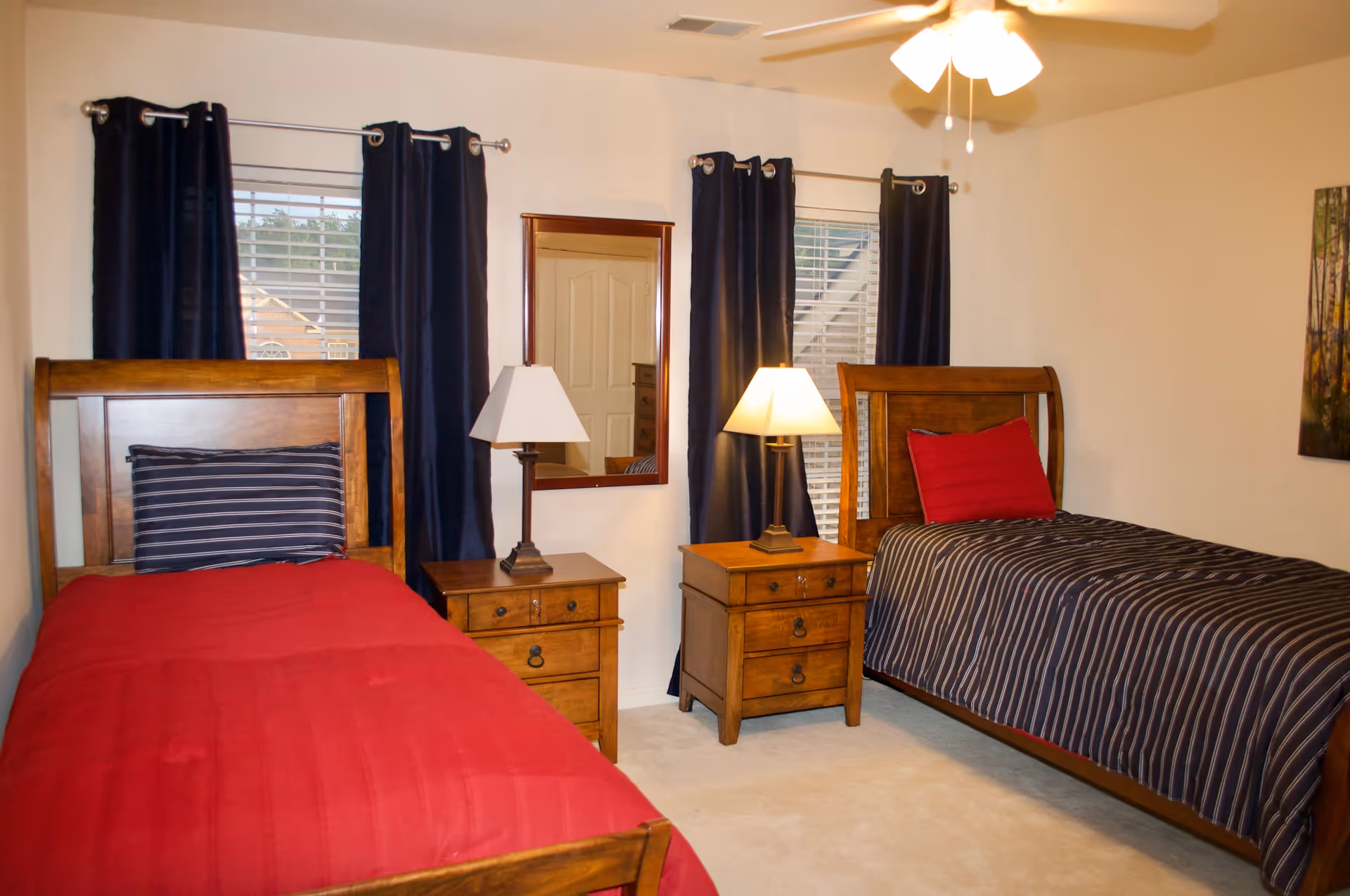 A bedroom with two twin beds featuring wooden frames, matching nightstands with lamps, dark curtains, and a ceiling fan.