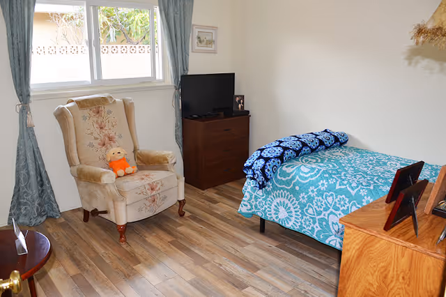 A cozy bedroom with a single bed covered in a blue and white floral patterned blanket and a folded blue blanket on top. Next to the bed is a wooden dresser with framed photos. Across from the bed is a beige upholstered armchair with floral patterns and an orange stuffed toy placed on it. A small dark wooden table is partially visible in the foreground. A window with blue curtains lets in natural light, and a small TV sits on a dark wooden chest of drawers against the wall.