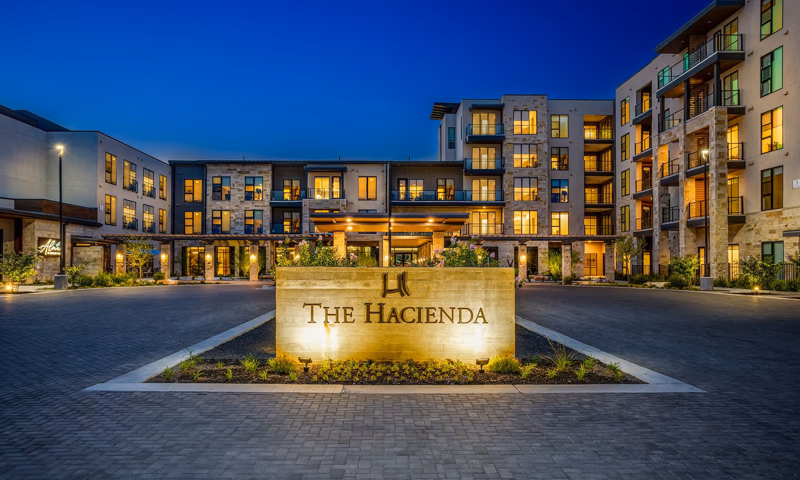 Exterior view of The Hacienda at Georgetown senior living facility at dusk, showing a modern multi-story building with illuminated windows and a large stone sign in the foreground with the facility's name.