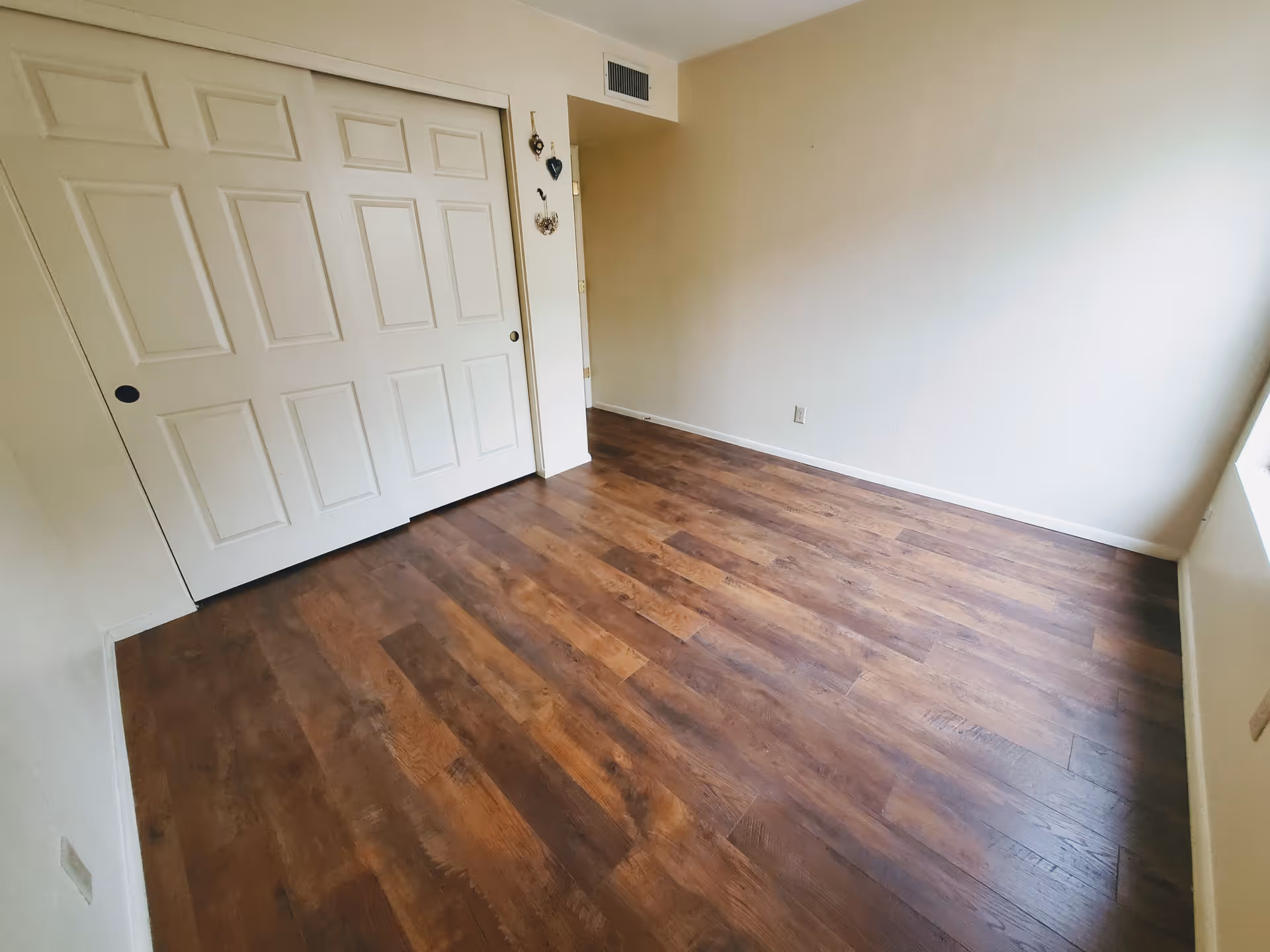 Empty room with wooden flooring, white walls, a large window on the right side, and a white sliding closet door on the left side.