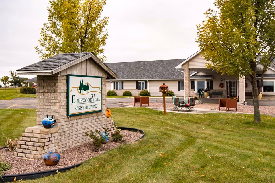 Exterior view of Edgewood Vista Assisted Living facility showing a brick sign with the facility name, a green lawn with some trees, a paved driveway, and a building with beige siding and a covered entrance with outdoor seating.