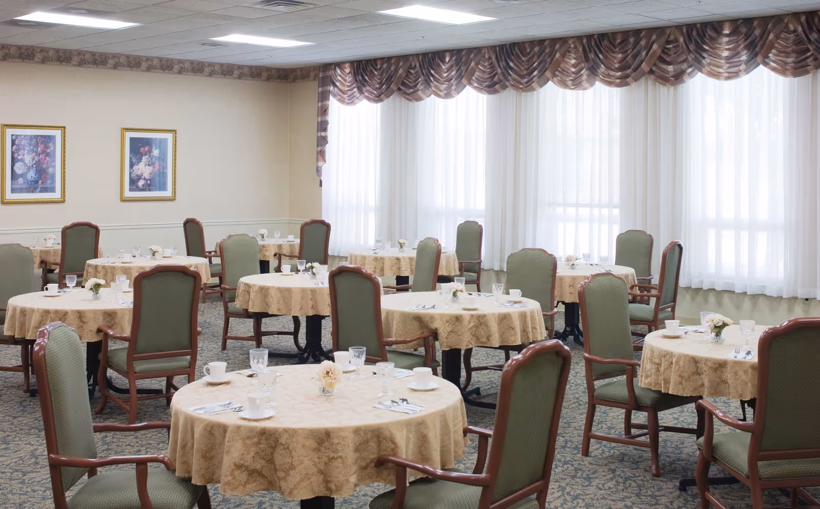 Bright dining room with round, cloth-covered tables set for meals and green upholstered chairs beneath large curtained windows.
