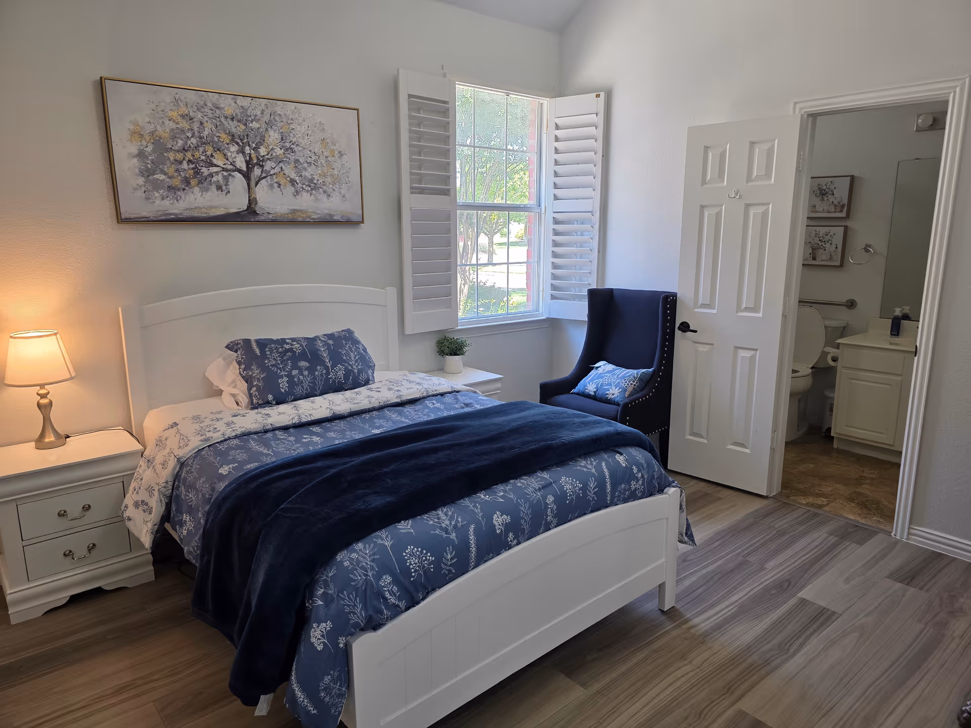 White bedroom with blue floral bedding, nightstands, lamp, window shutters, and open bathroom door.