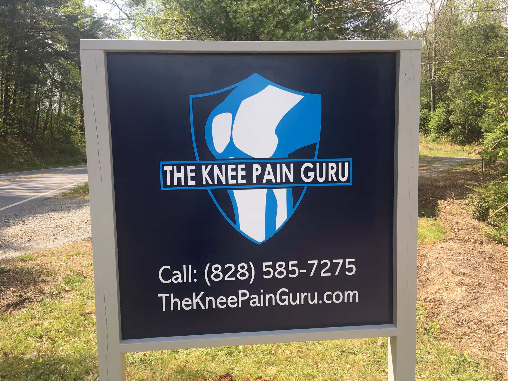 Outdoor signboard featuring The Knee Pain Guru logo and contact information in a wooded area.