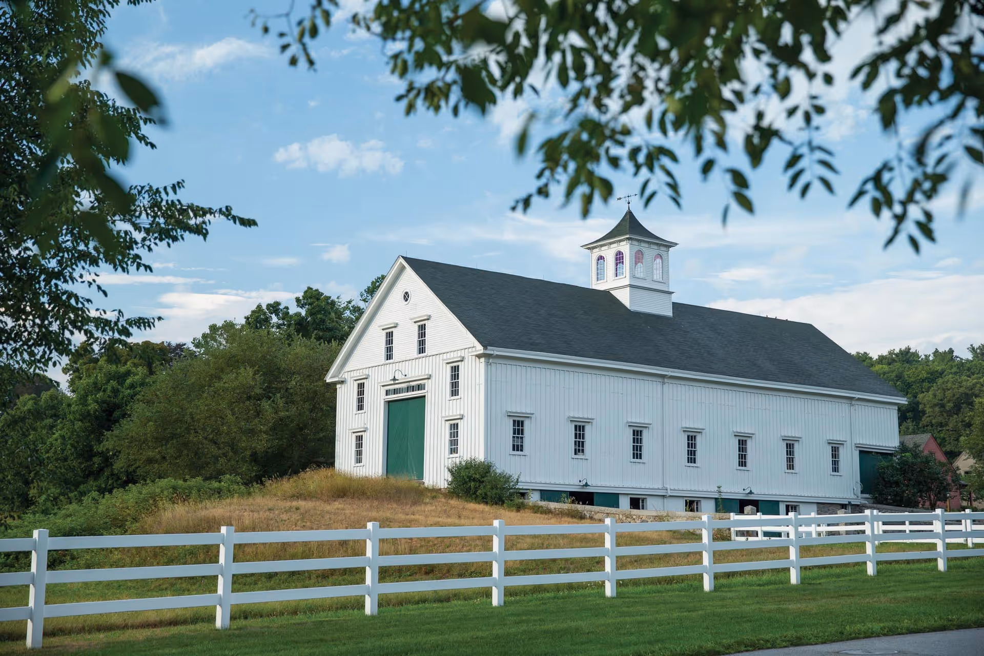 A large white barn-style building with a dark roof and a small cupola on top, surrounded by green grass and trees, with a white wooden fence in the foreground under a partly cloudy blue sky.