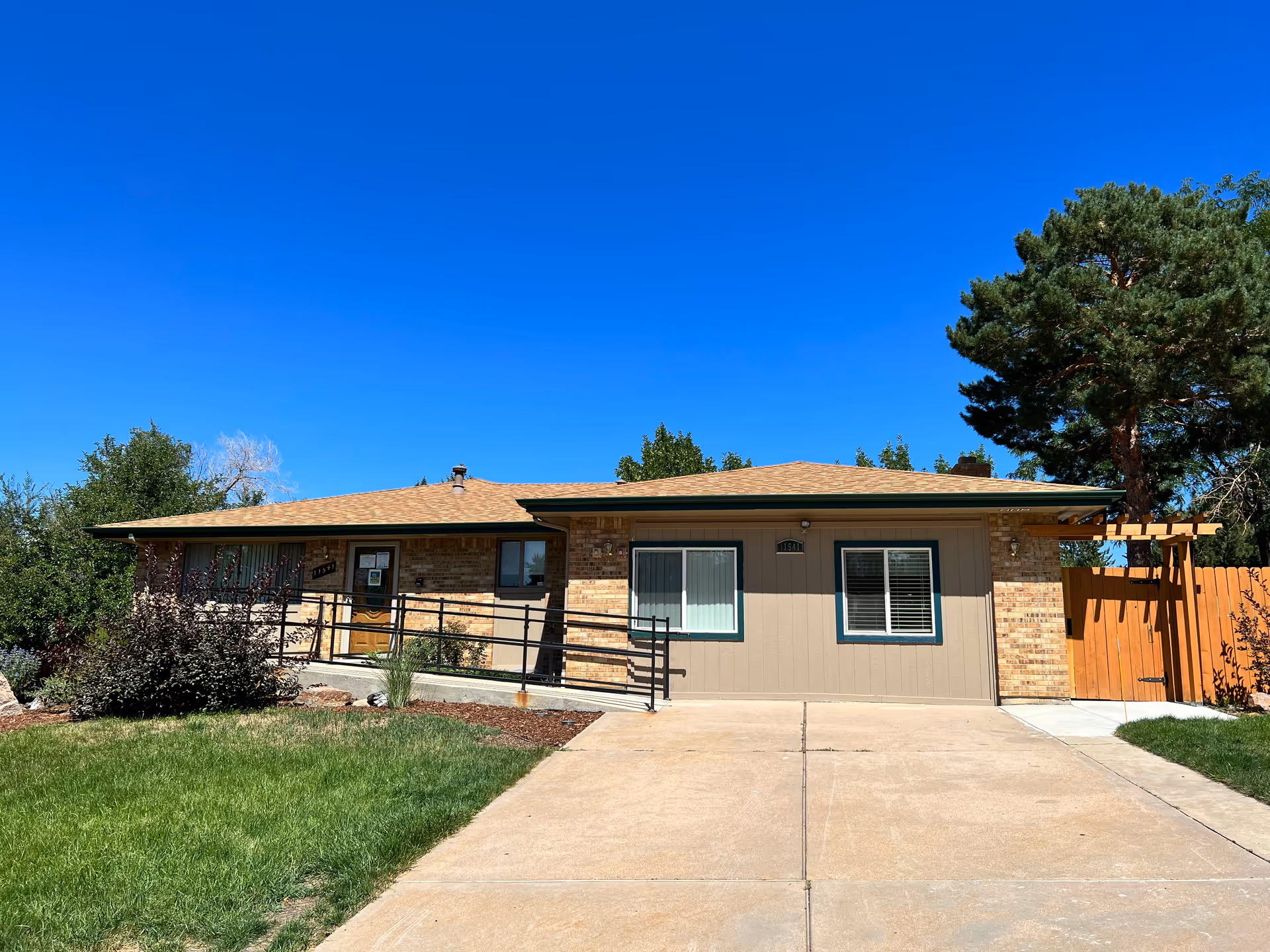 Single-story building with a beige and brick exterior under a clear blue sky. There is a concrete driveway leading up to the entrance, which has a ramp with black railings. The building is surrounded by green grass, bushes, and trees.
