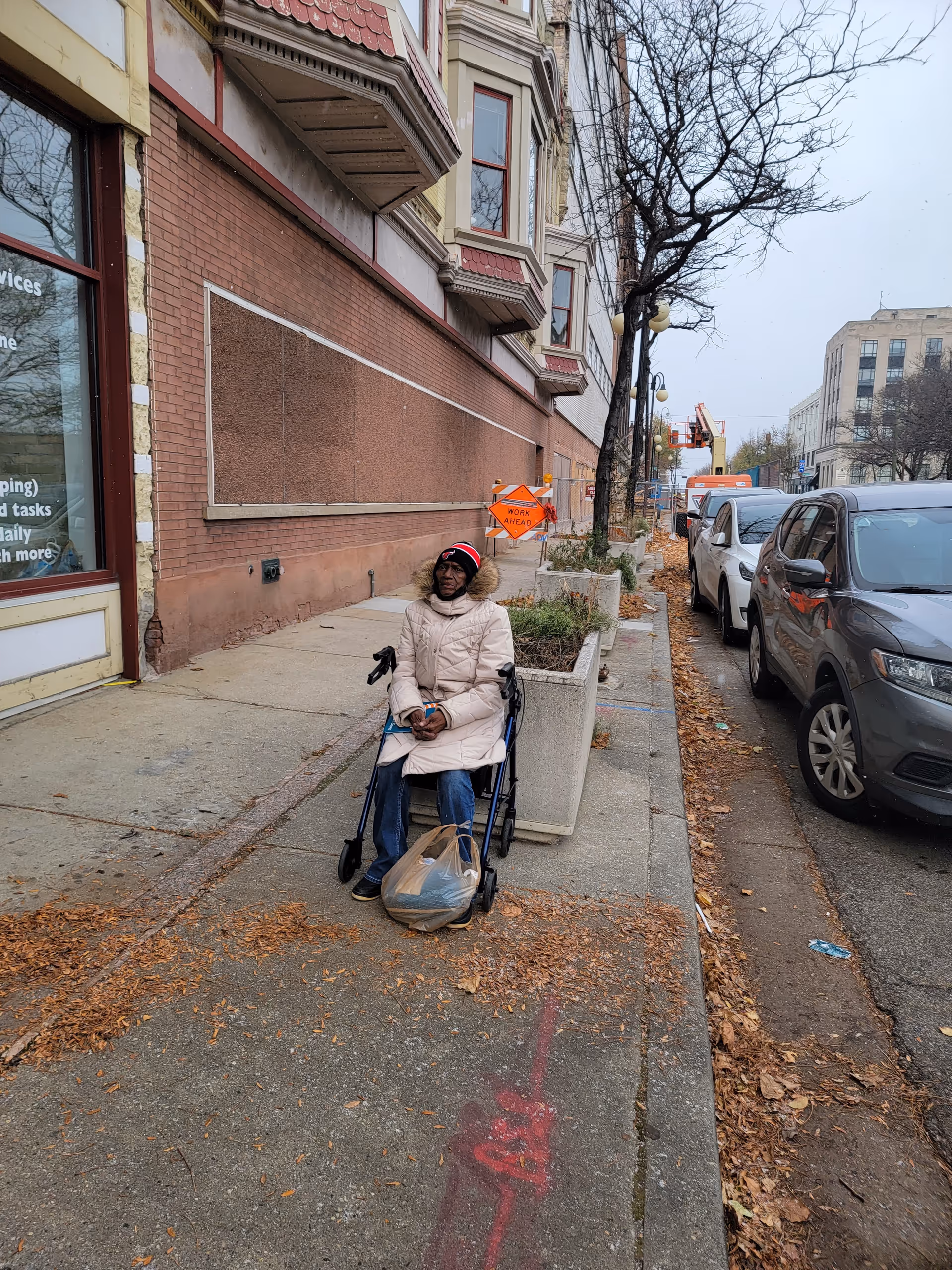 Person in a beige coat sits in a wheelchair on a city sidewalk.