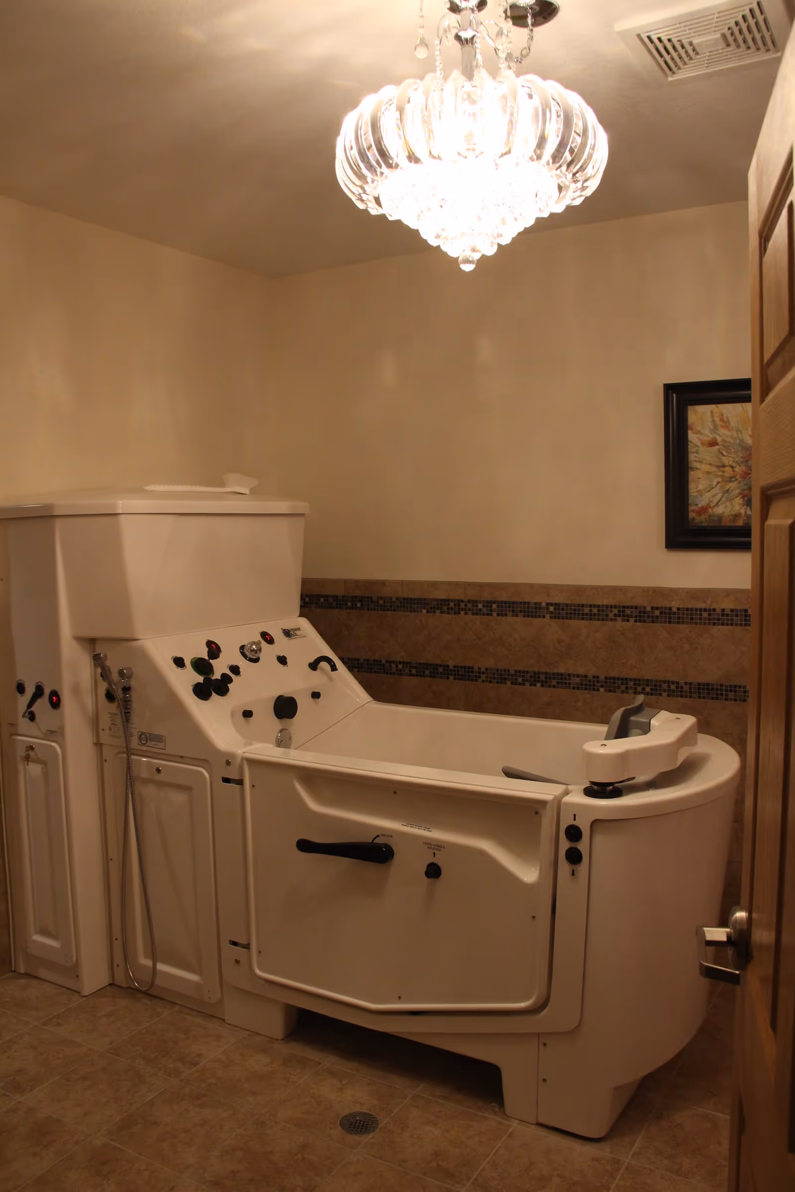 A walk-in bathtub with control knobs and a handheld showerhead in a bathroom with beige tiled floor and walls, a wooden door, a framed picture on the wall, and a decorative ceiling light fixture.