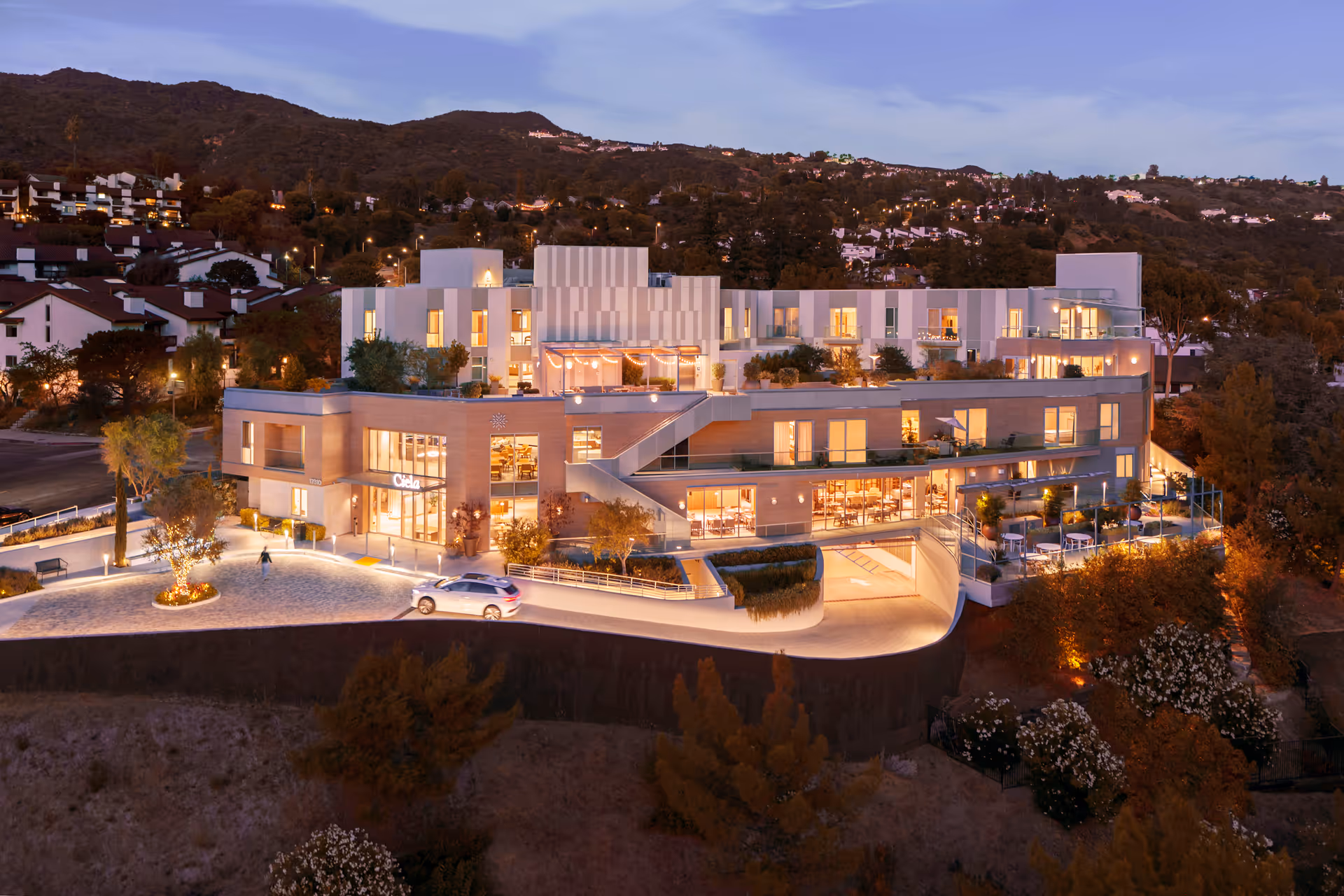 Evening view of Ciela Senior Living facility situated on a hillside with warm interior lights glowing through large windows. The modern multi-story building features outdoor seating areas, a driveway with a car, and surrounding trees and hills in the background.