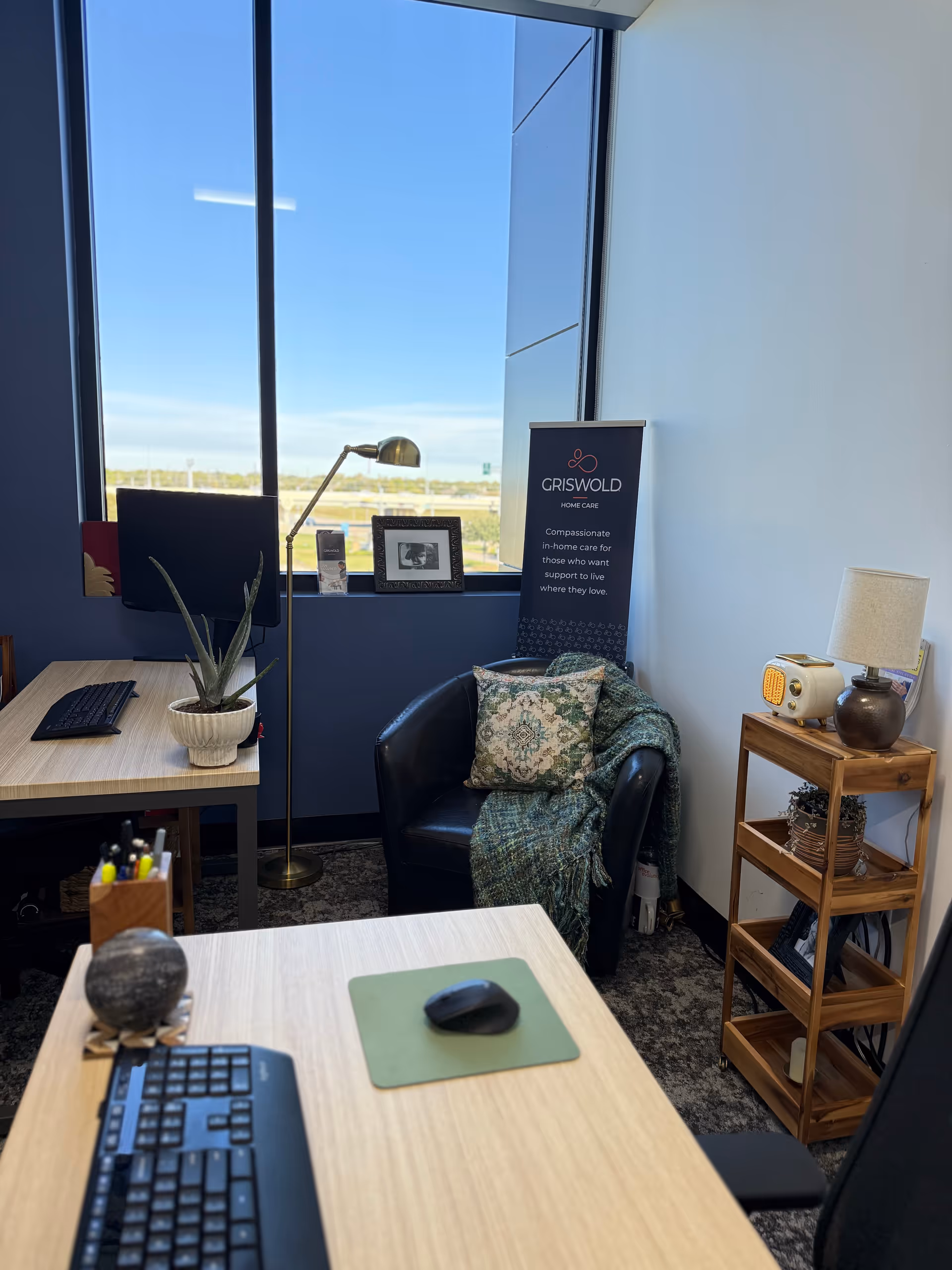 Office space with desks, a computer, plant, and Griswold banner by a large window.