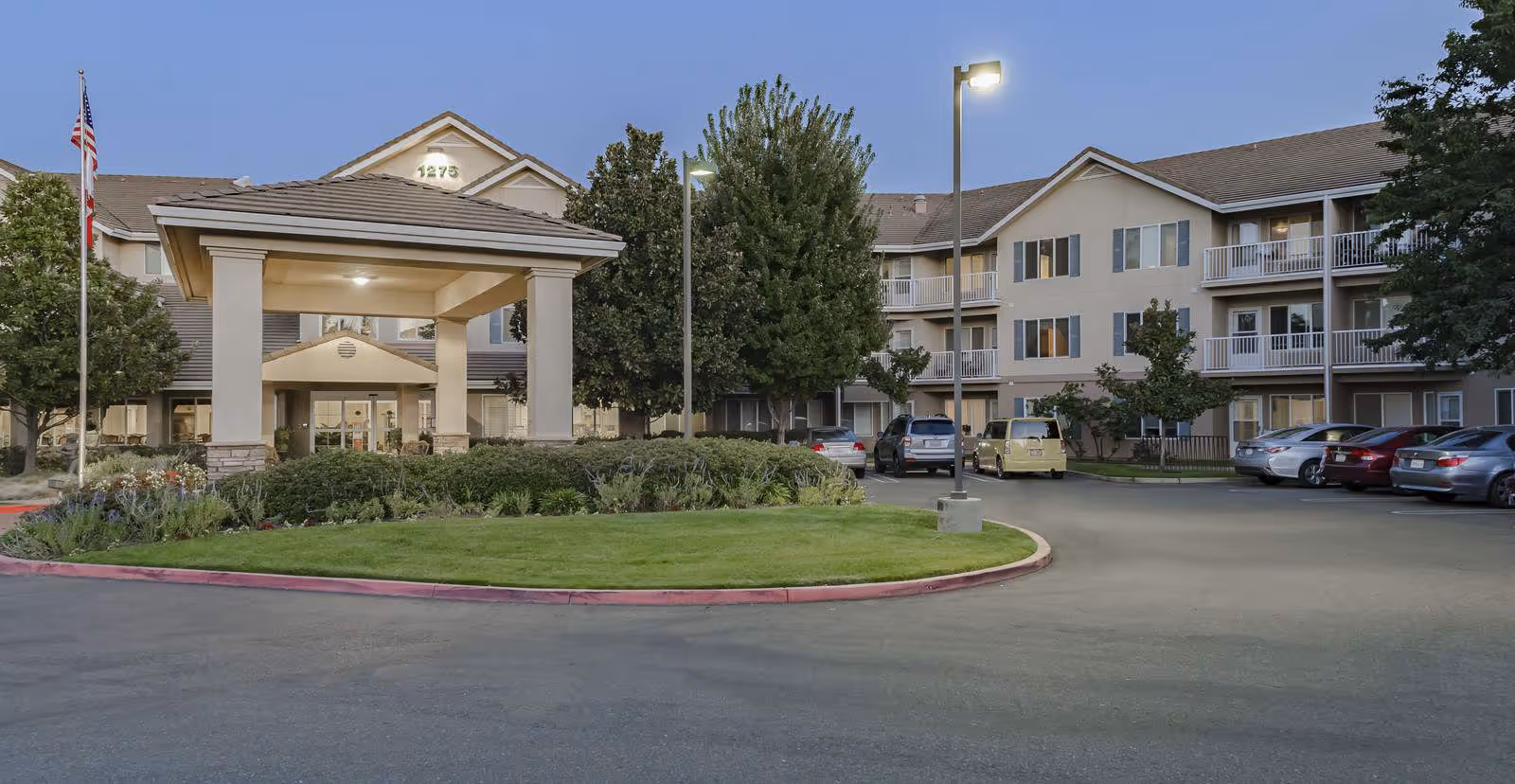 Exterior view of Truewood by Merrill, Roseville senior living facility at dusk, showing a three-story building with balconies, a covered entrance with pillars, landscaped greenery, parked cars, and street lamps.