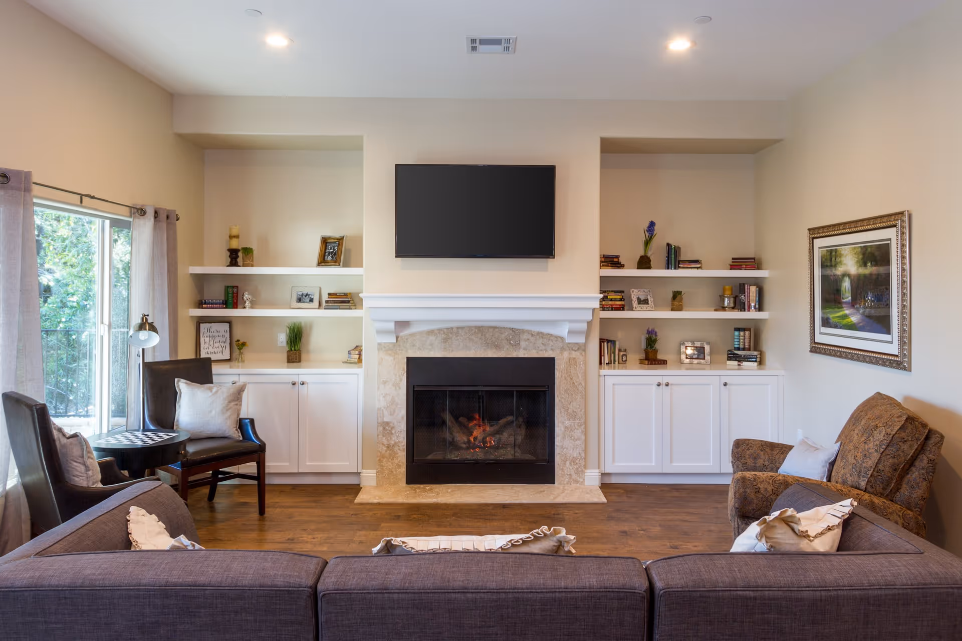 A cozy living room with a gray sofa facing a fireplace with a mounted flat-screen TV above it. On either side of the fireplace are built-in white shelves and cabinets decorated with books, framed photos, candles, and small plants. To the left, there are two dark chairs with a small round table between them near a large window with light curtains. To the right, there is a patterned armchair and a framed picture on the wall. The room has wooden flooring and recessed ceiling lights.