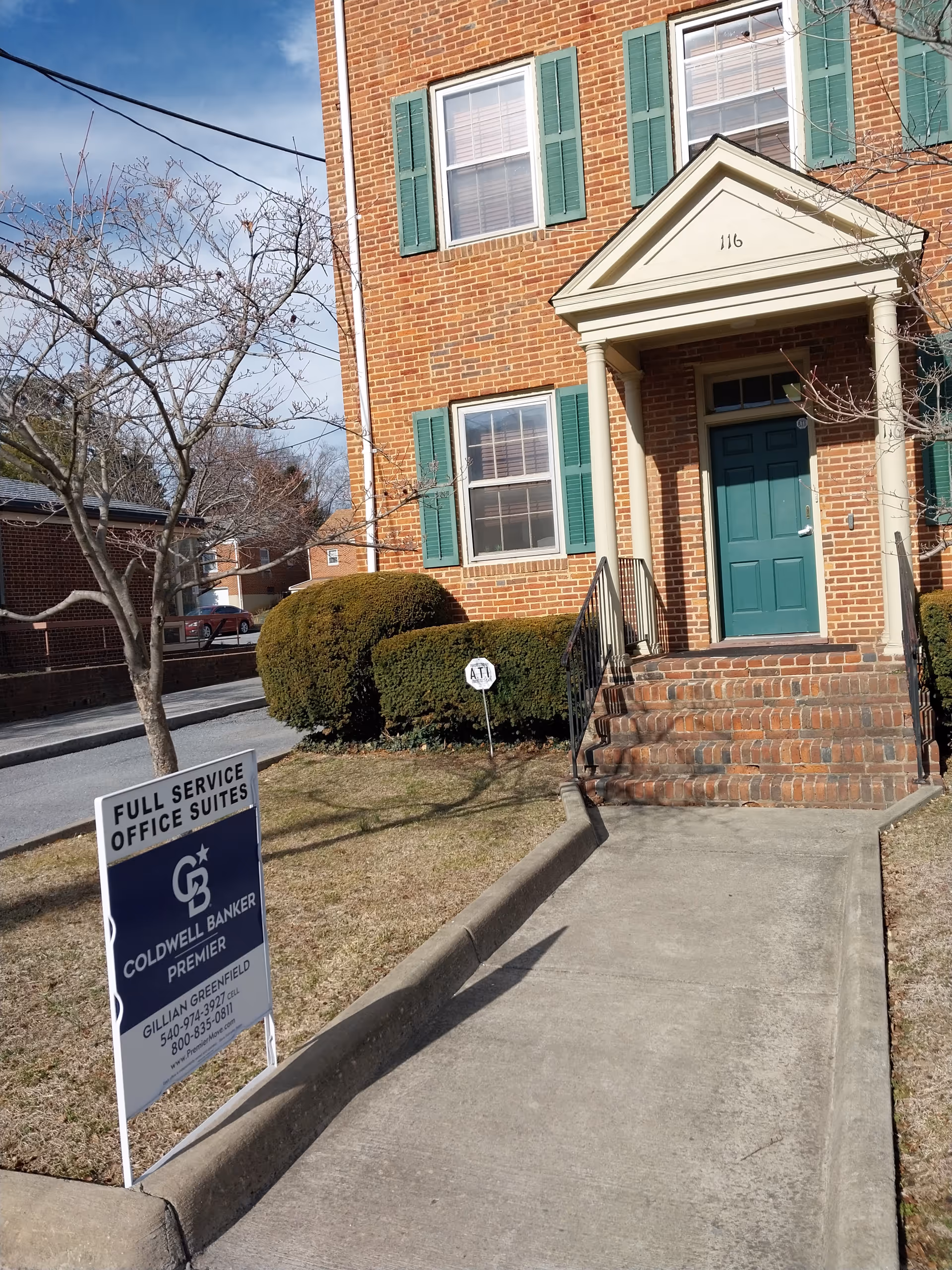 Brick building entrance with teal door, green shutters, stairs, and a lawn sign.