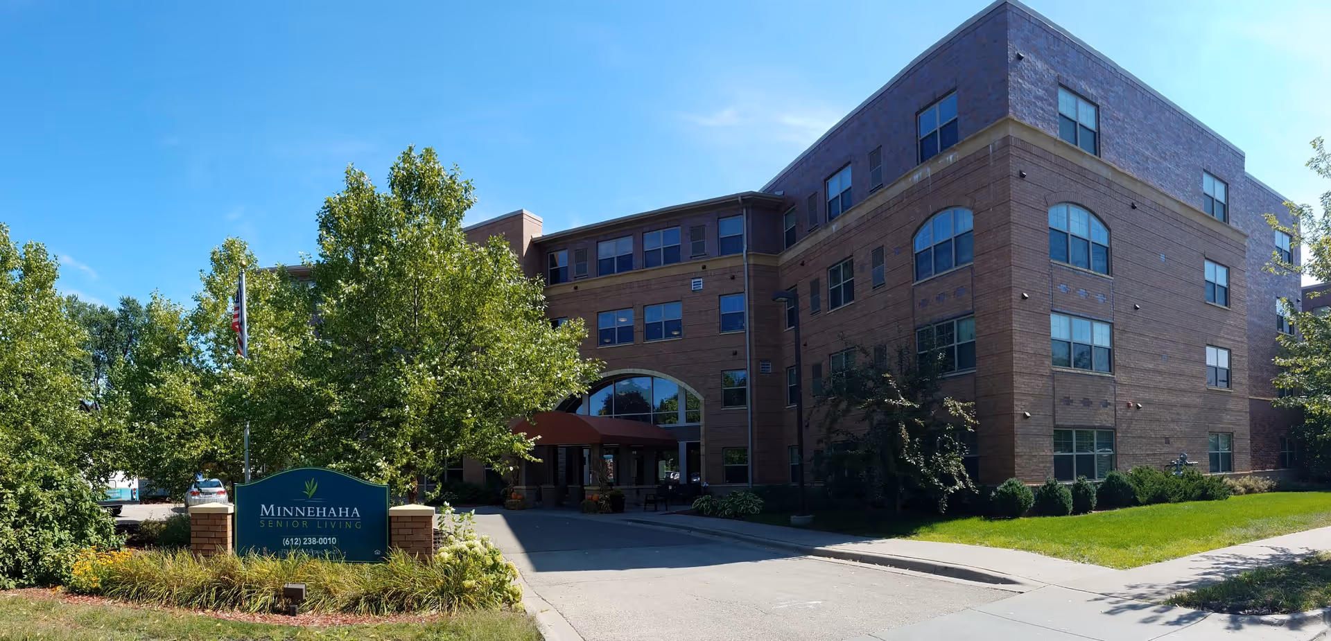 Brick senior living building with entrance, trees, and landscaped sign at driveway.