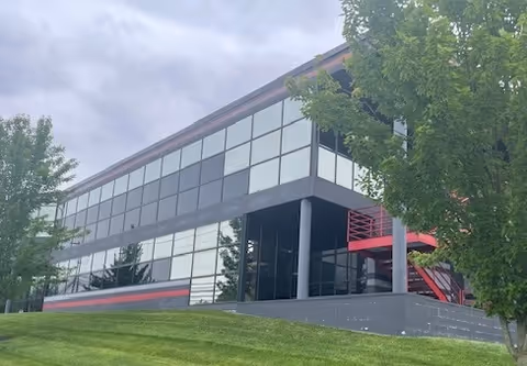 Modern glass-front exterior of a building with red accents, stairs, trees, and cloudy sky.
