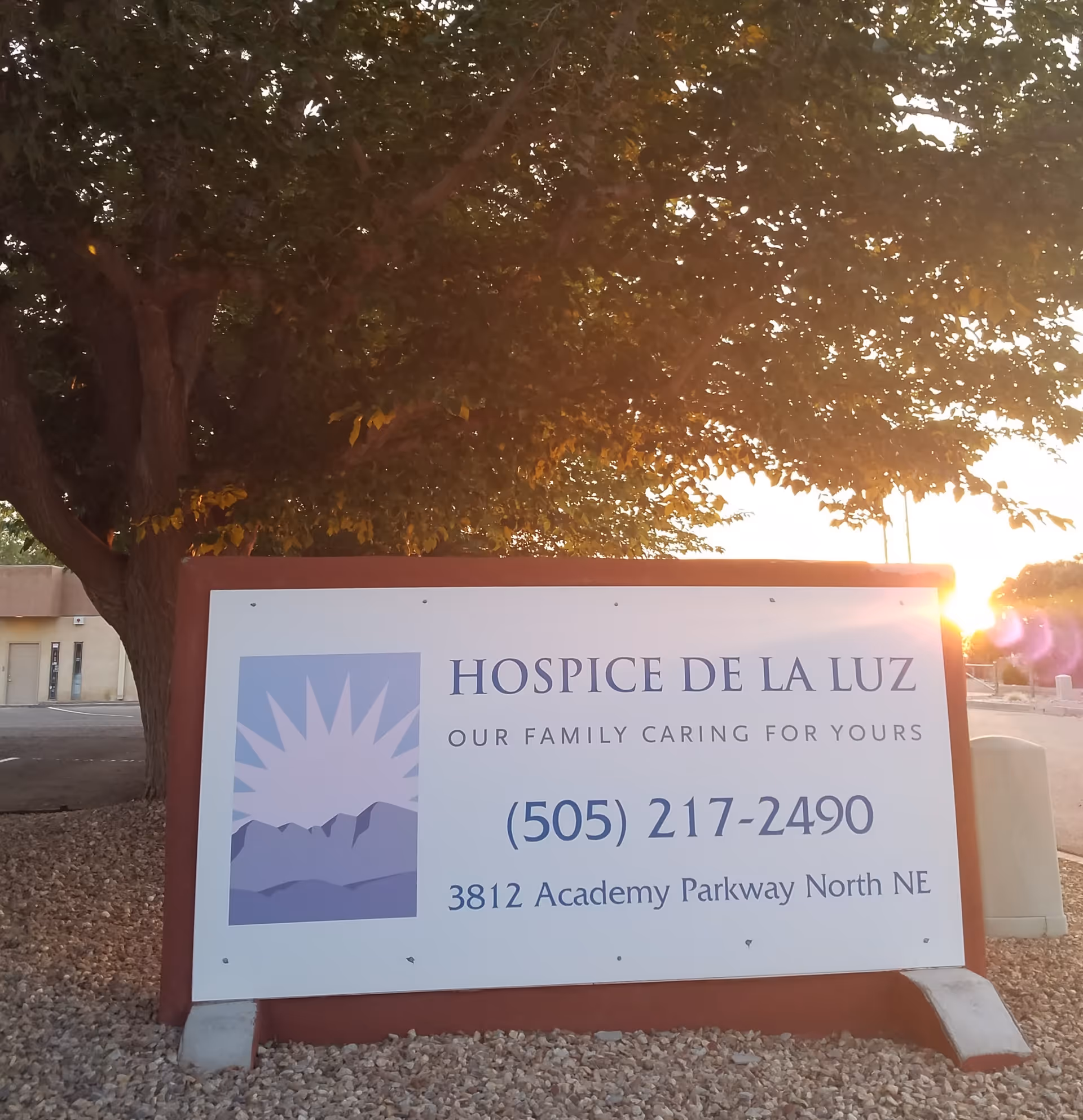 Outdoor sign for Hospice De La Luz with phone number, address, and a sunlit backdrop.