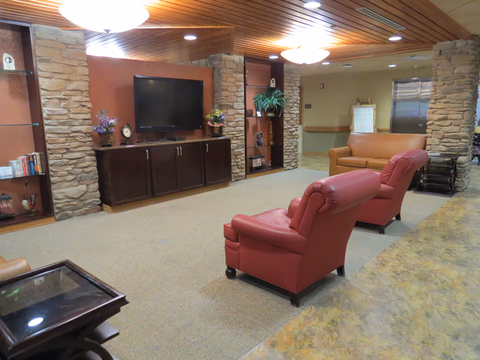 A cozy living room area with two red leather armchairs and a brown leather sofa arranged around a wall-mounted flat-screen TV. The TV is mounted on a reddish-brown accent wall with stone pillars on either side. There are shelves with decorative items and books, a glass-top side table, and a ceiling with wooden paneling and recessed lighting. The floor is carpeted, and there is a hallway with an elevator visible in the background.