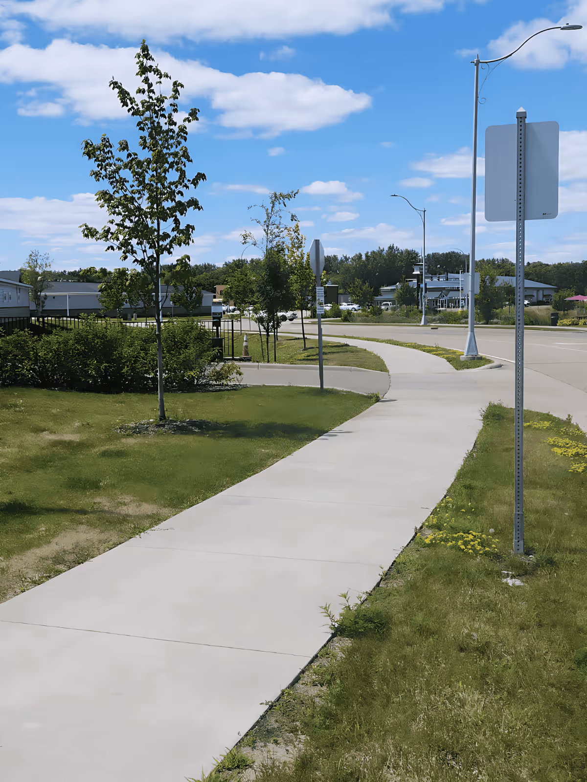 Wide concrete sidewalk curves through grassy outdoor area with trees and street signs.