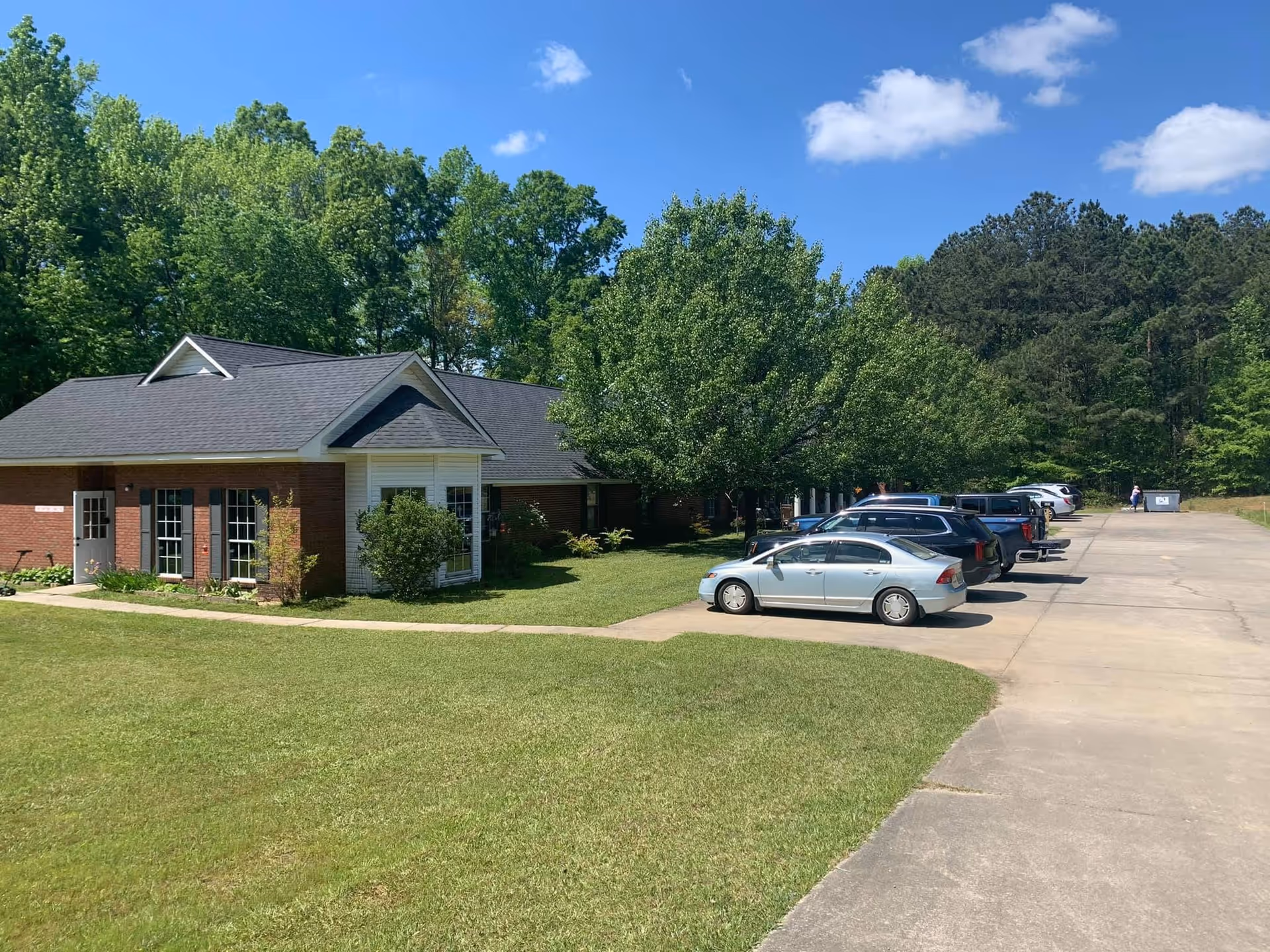 Front view of a single-story brick assisted living building with a parking lot and trees.