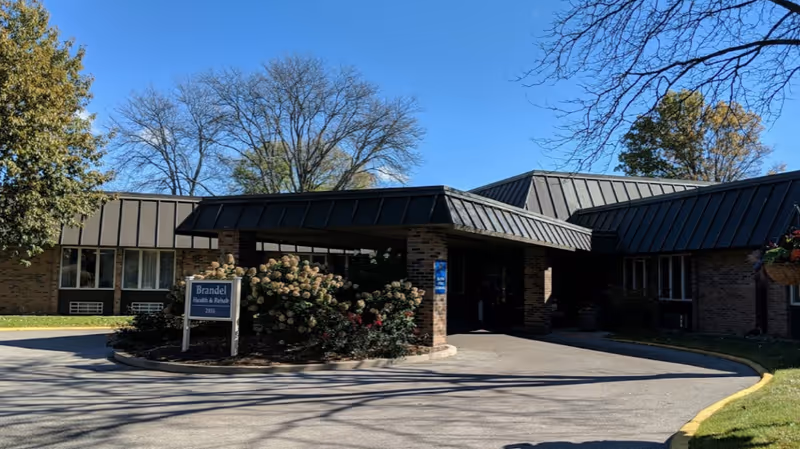 Front entrance of Brandel Care Center showing a covered drop-off, sign, and landscaped shrubs.
