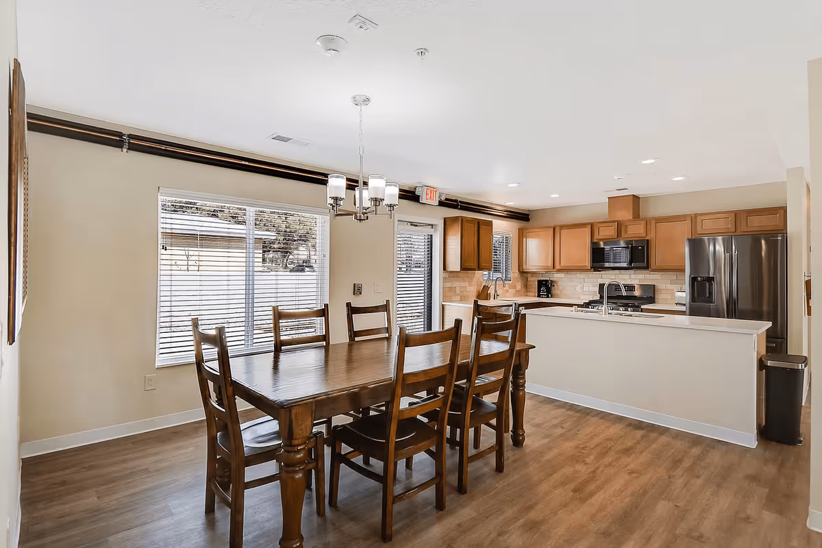 A bright and spacious dining area with a wooden dining table and six matching chairs. The room features large windows with blinds, a modern chandelier above the table, and an open kitchen with wooden cabinets, stainless steel appliances, and a white countertop. The floor is covered with light wood laminate.