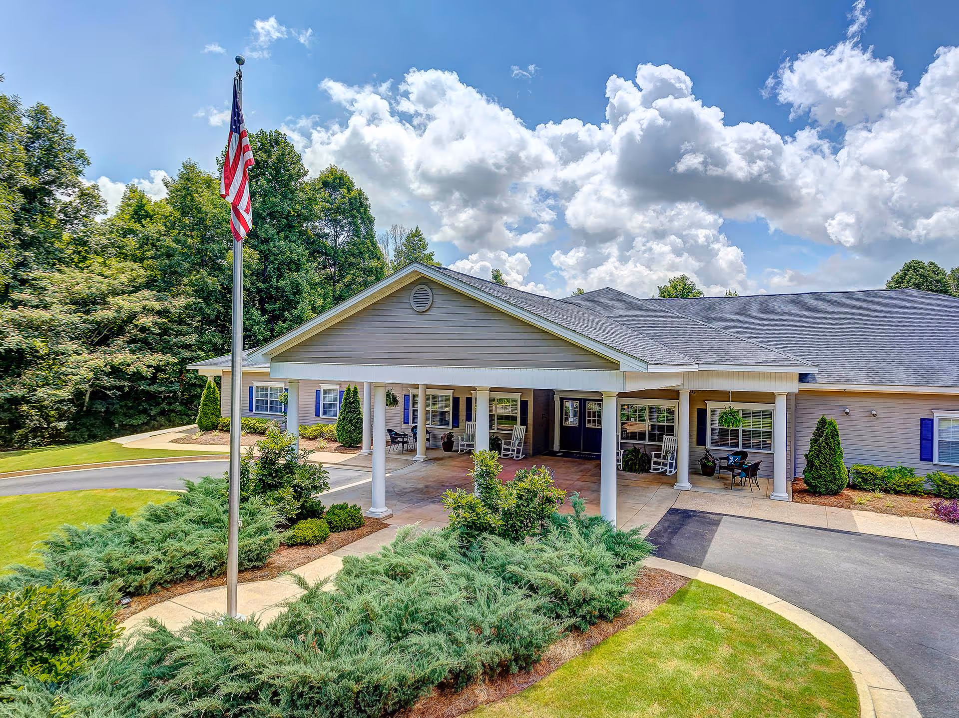 Single-story senior living facility entrance with covered porch, flagpole, and landscaped driveway.