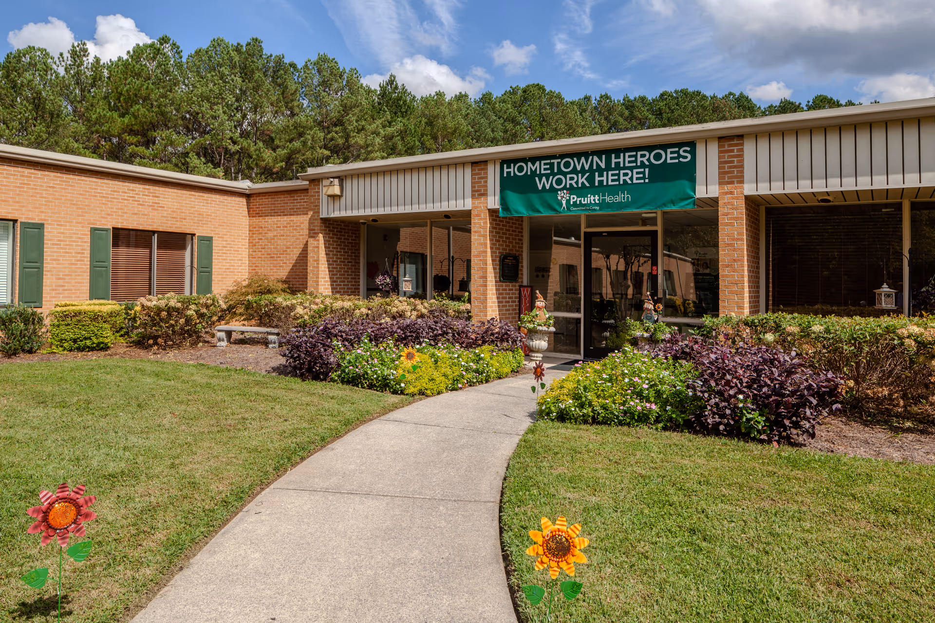 Brick senior living facility entrance with a curved walkway and flower beds under a green banner.