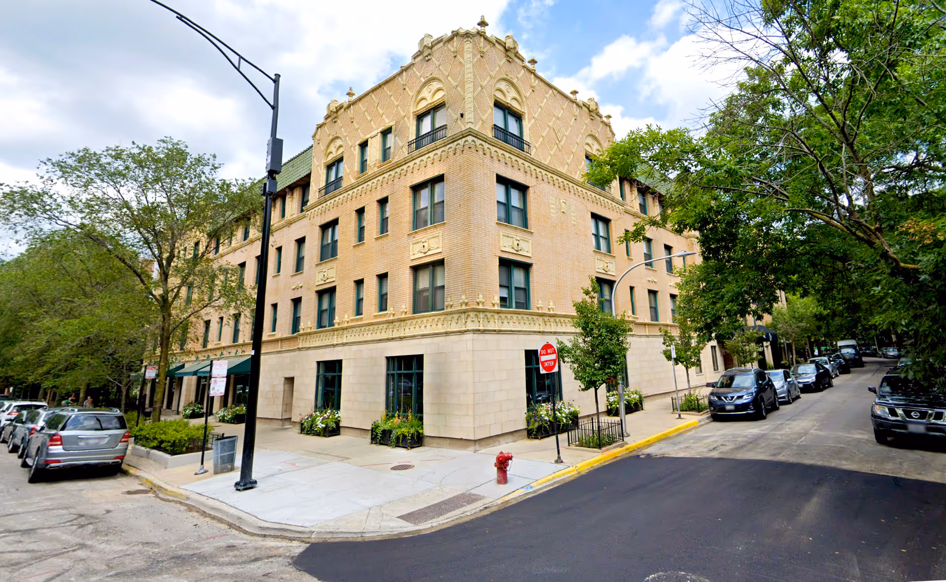 Corner view of a multi-story beige brick building with decorative architectural details, surrounded by trees and parked cars along the streets on both sides. The sky is partly cloudy.