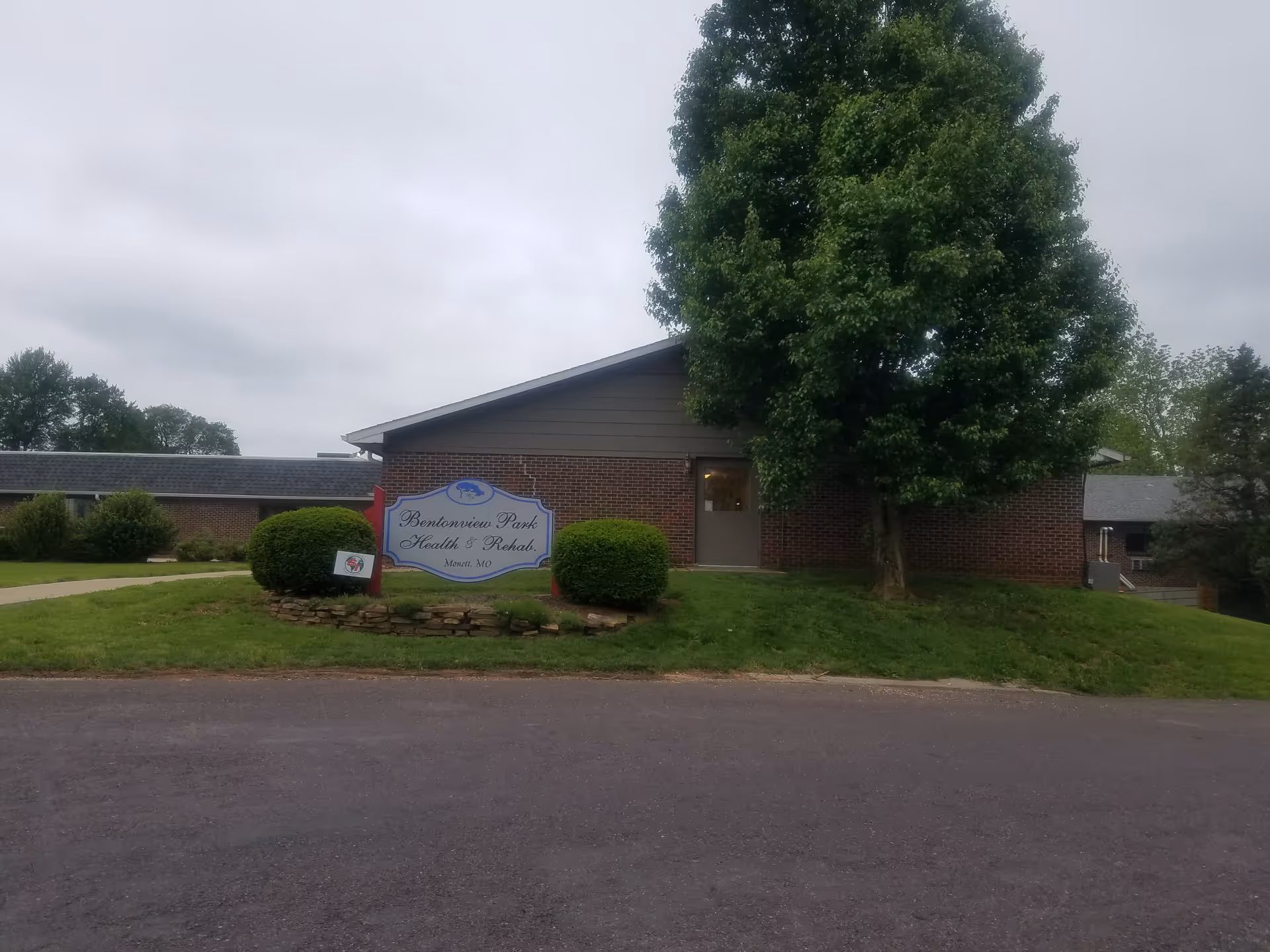 Brick facility exterior with Bentonview Park Health & Rehab sign, front lawn, and a large tree.