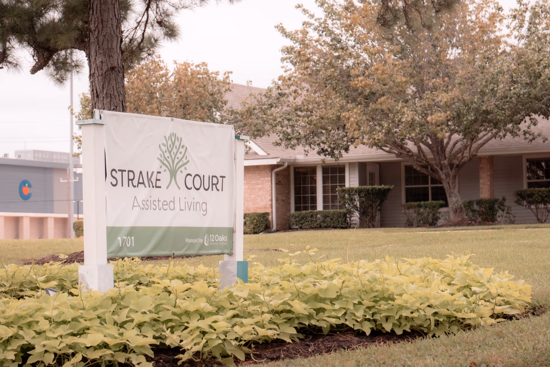 A yard sign that reads "Strake Court Assisted Living" in front of a single-story building with trees and landscaping.