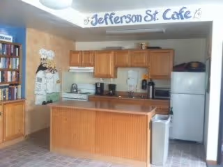 Communal kitchenette with wooden cabinets, a center island, refrigerator and microwave, a coffee station and bookshelf under a 'Jefferson St. Cafe' sign.