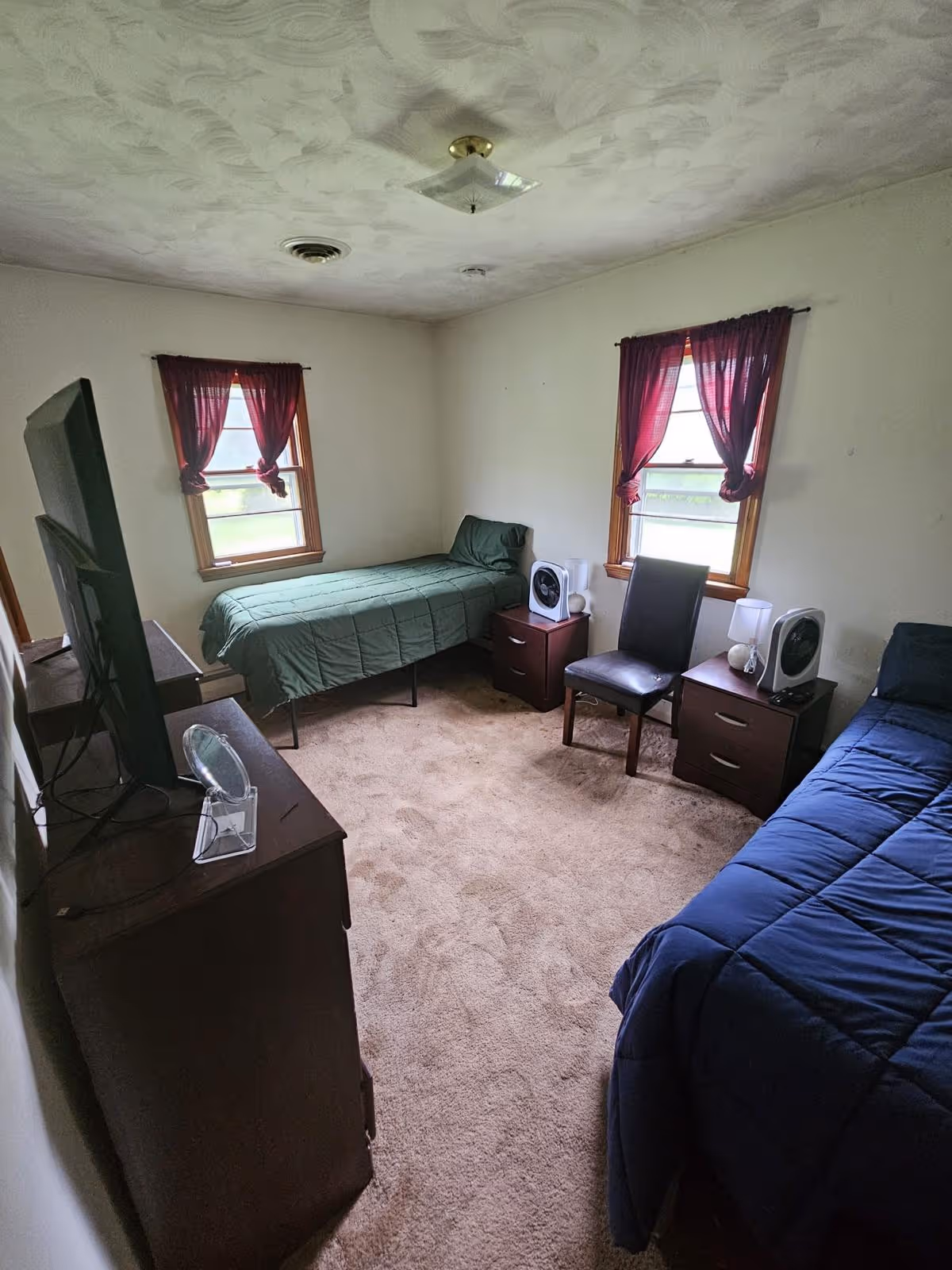 A bedroom with two beds, a dresser, TV, and two nightstands by windows with red curtains.