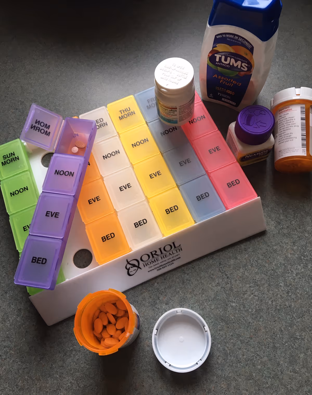 A colorful weekly medication organizer with pills, bottles, and Oriol Home Health branding on a counter.