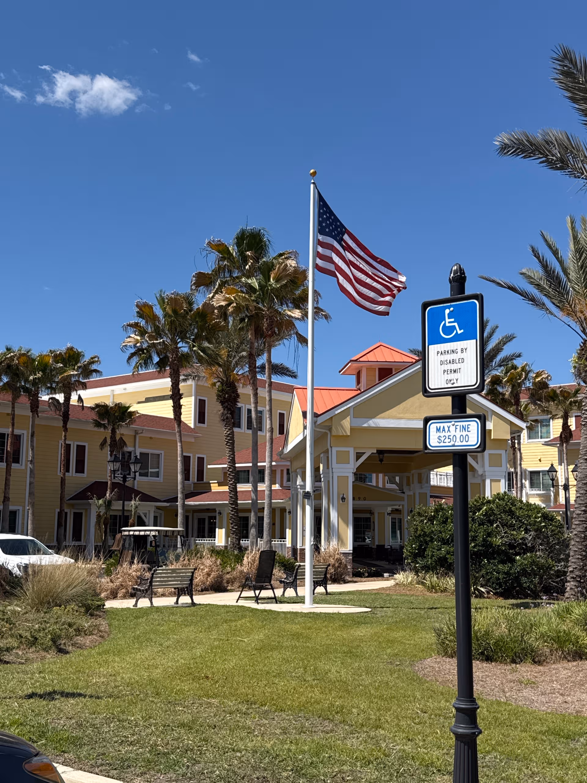 Exterior view of Buffalo Crossings Assisted Living with flag, palm trees, benches, and a sunny sky.