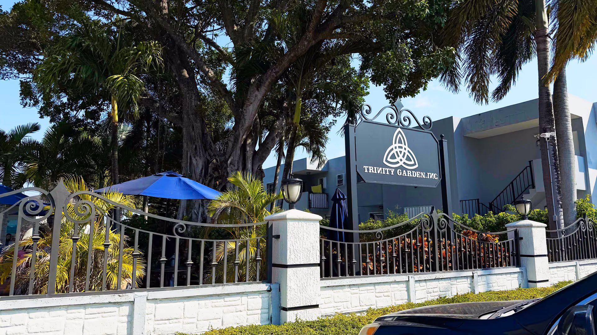 Exterior view of a gated senior living entrance with a sign and umbrellas.