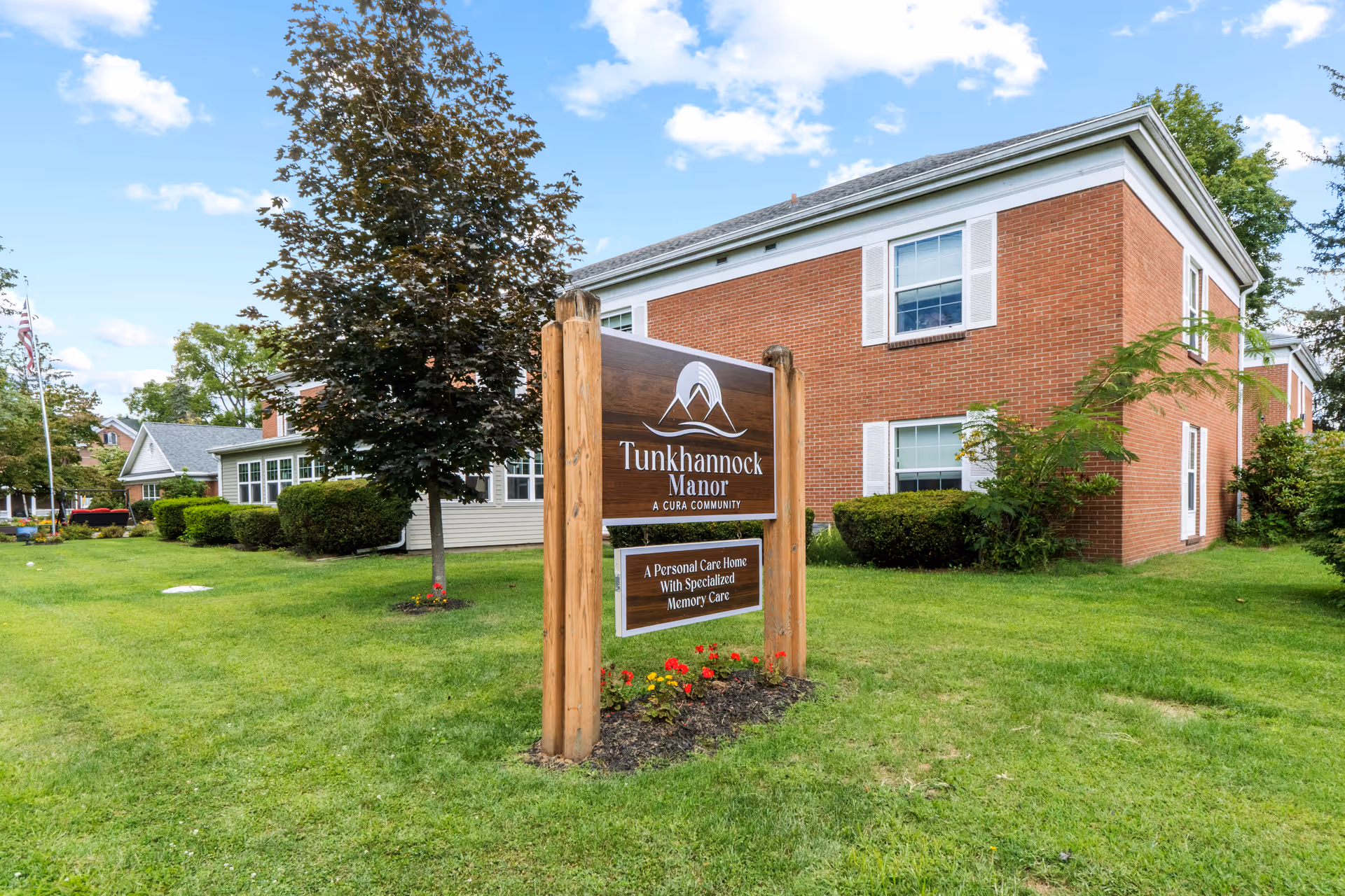 Brick senior living facility with a front lawn, trees, and a wooden sign reading Tunkhannock Manor.