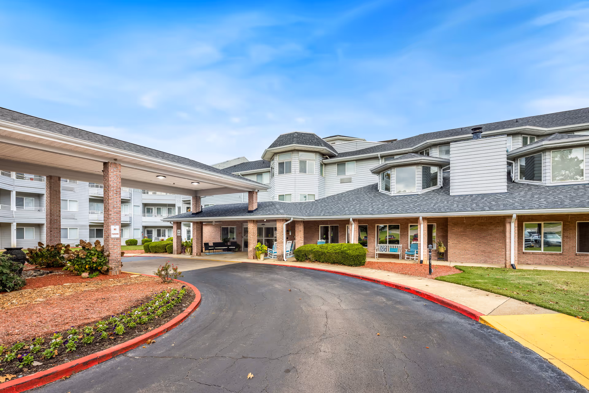 Exterior view of Jackson Meadow - A Provincial Senior Living Community showing a curved driveway leading to a covered entrance. The building has multiple windows, brick and white siding, and a well-maintained landscape with shrubs and flower beds under a partly cloudy blue sky.