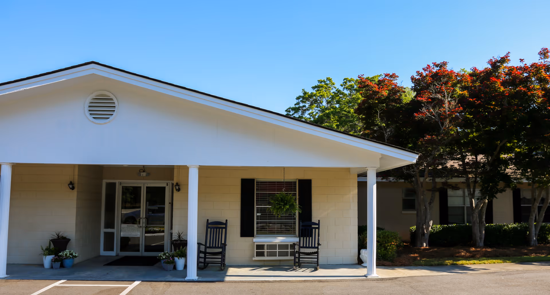 Exterior view of a single-story building with a covered entrance supported by white columns. Two black rocking chairs and potted plants are placed near the entrance. Trees with green and red leaves are visible on the right side under a clear blue sky.