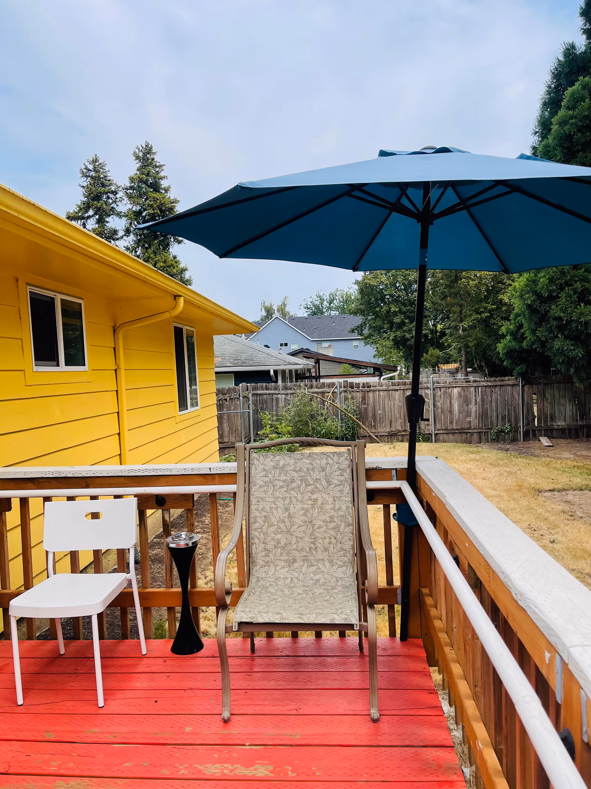 Outdoor patio area with a red wooden deck, a white chair, a beige patterned chair, and a large blue umbrella. The patio is adjacent to a yellow building with windows, and there is a wooden fence and trees in the background.