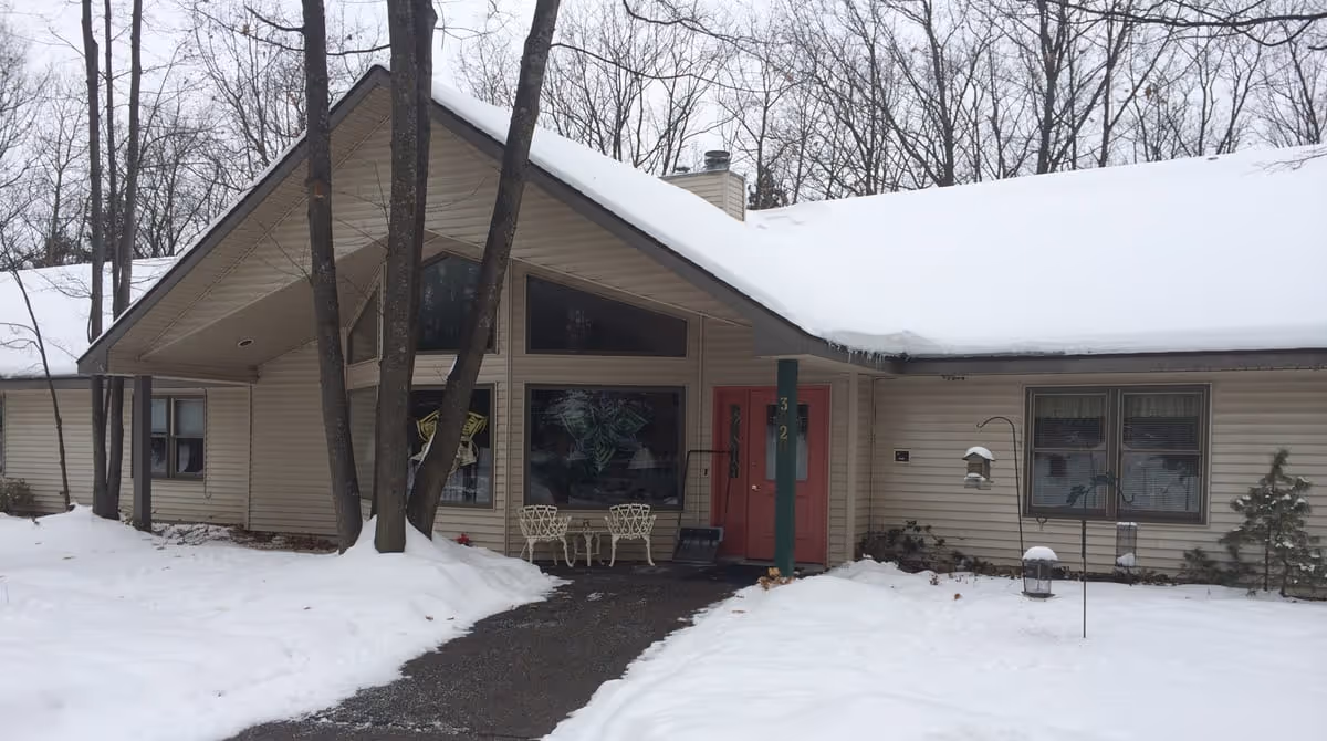 Snow-covered front entrance of a single-story assisted living building with a red door, patio chairs, and trees.