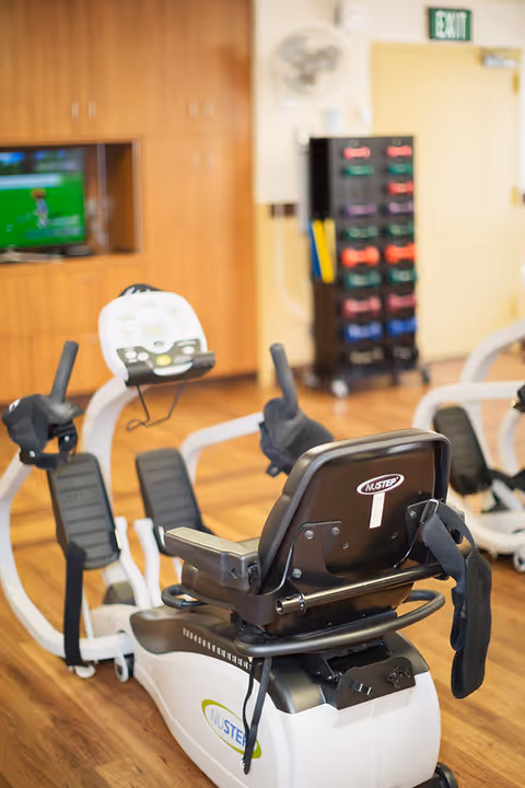 Recumbent exercise bikes and a rack of colorful dumbbells in a fitness room with wood floors and a TV.